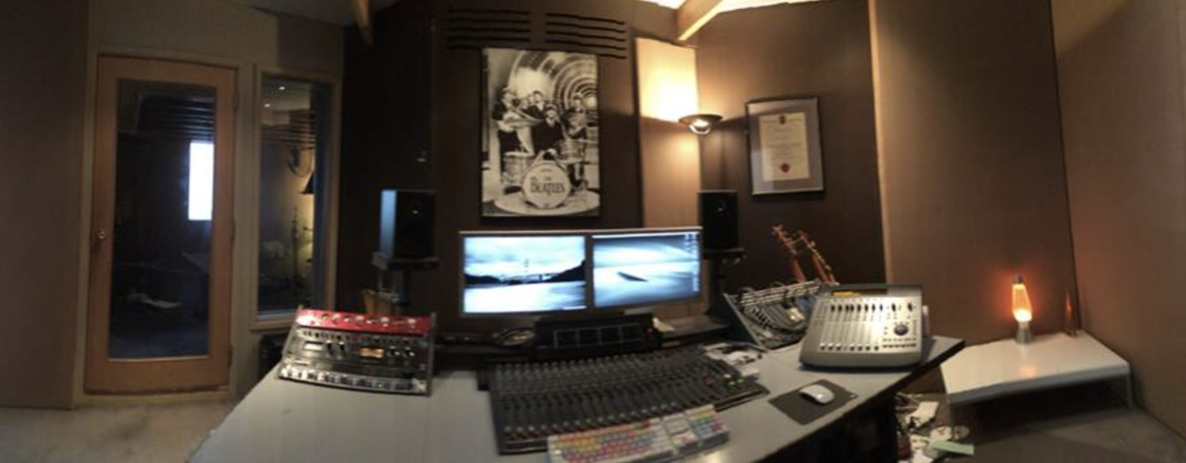 A recording studio features a control desk with multiple audio equipment, two computer monitors, and guitars on display, surrounded by acoustic treatment on the walls.