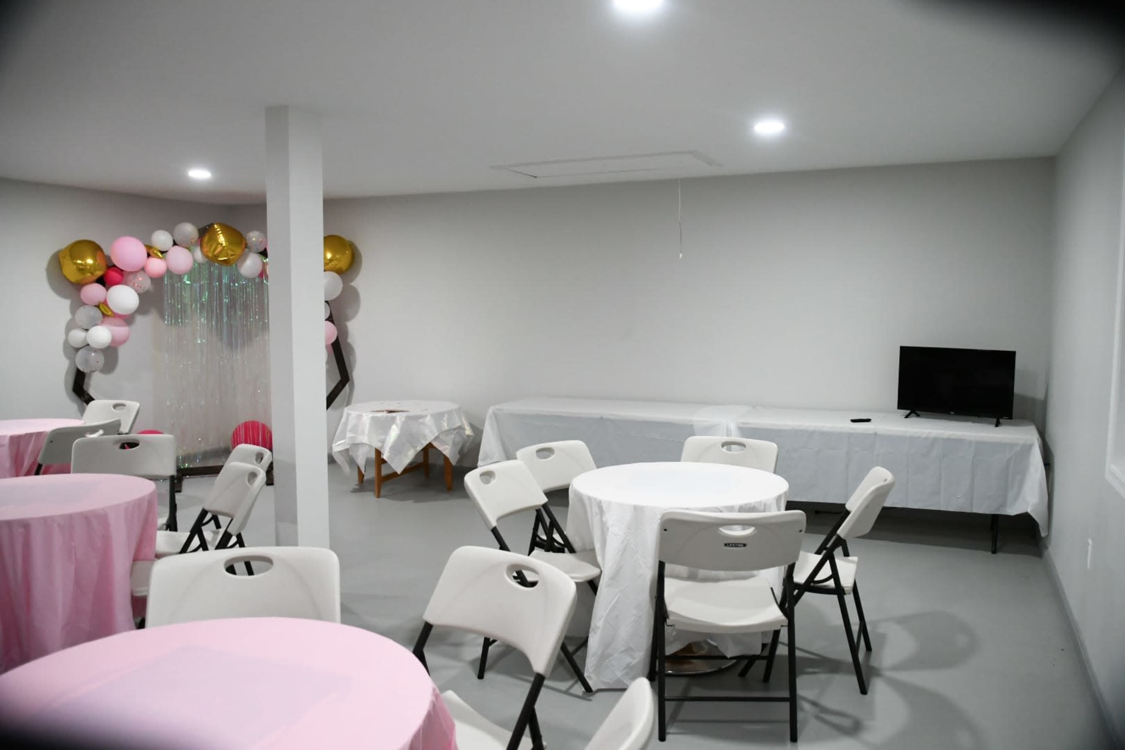 A spacious event room features round tables with pink tablecloths, white chairs, and a decorative backdrop of balloons against a plain wall.