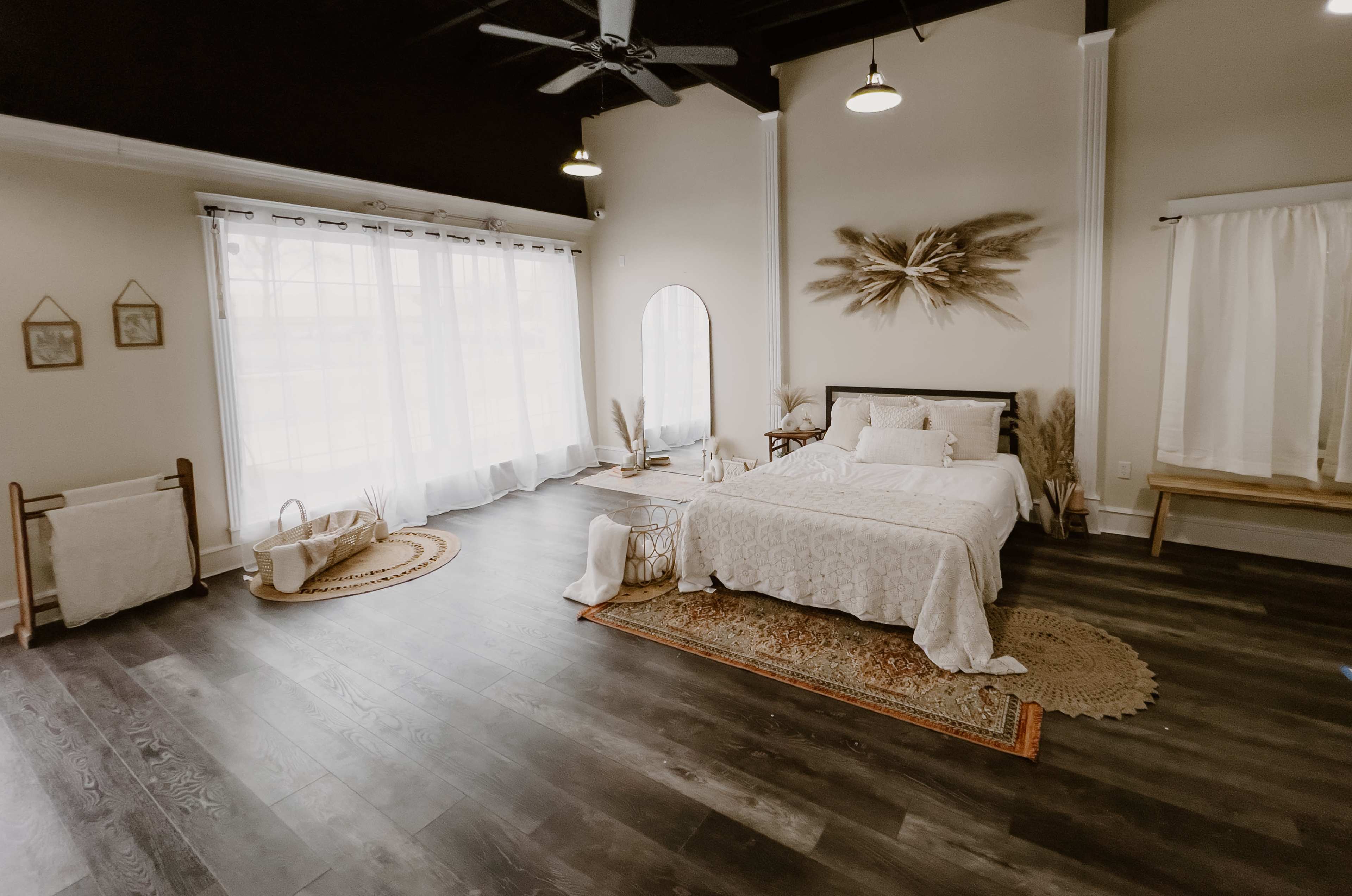 The image shows a well-lit bedroom with a bed, white bedding, large windows, and decorative elements like a mirror and woven baskets.
