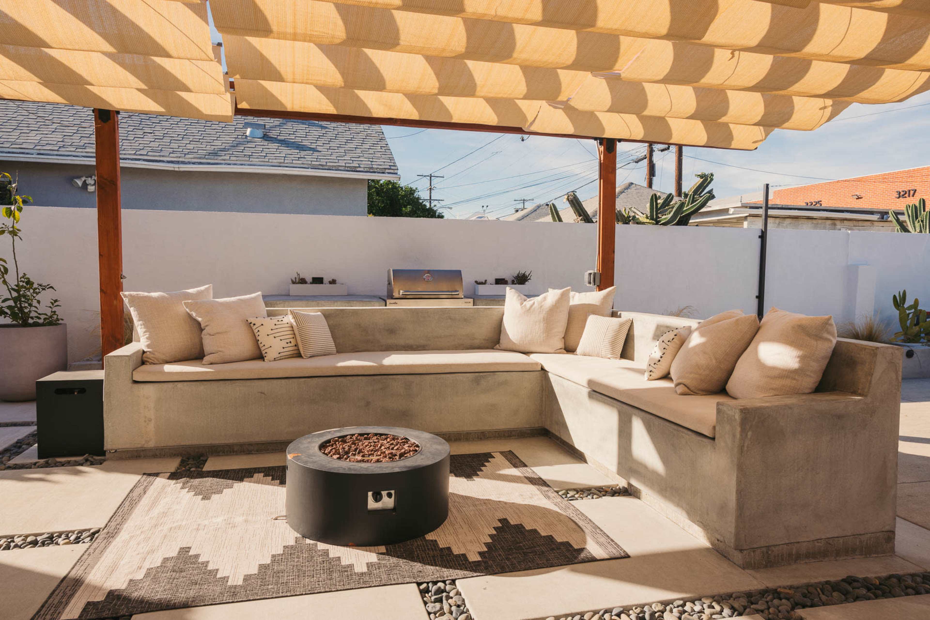 The scene features a modern outdoor seating area with a concrete sectional couch, a fire pit, and a large shaded pergola.