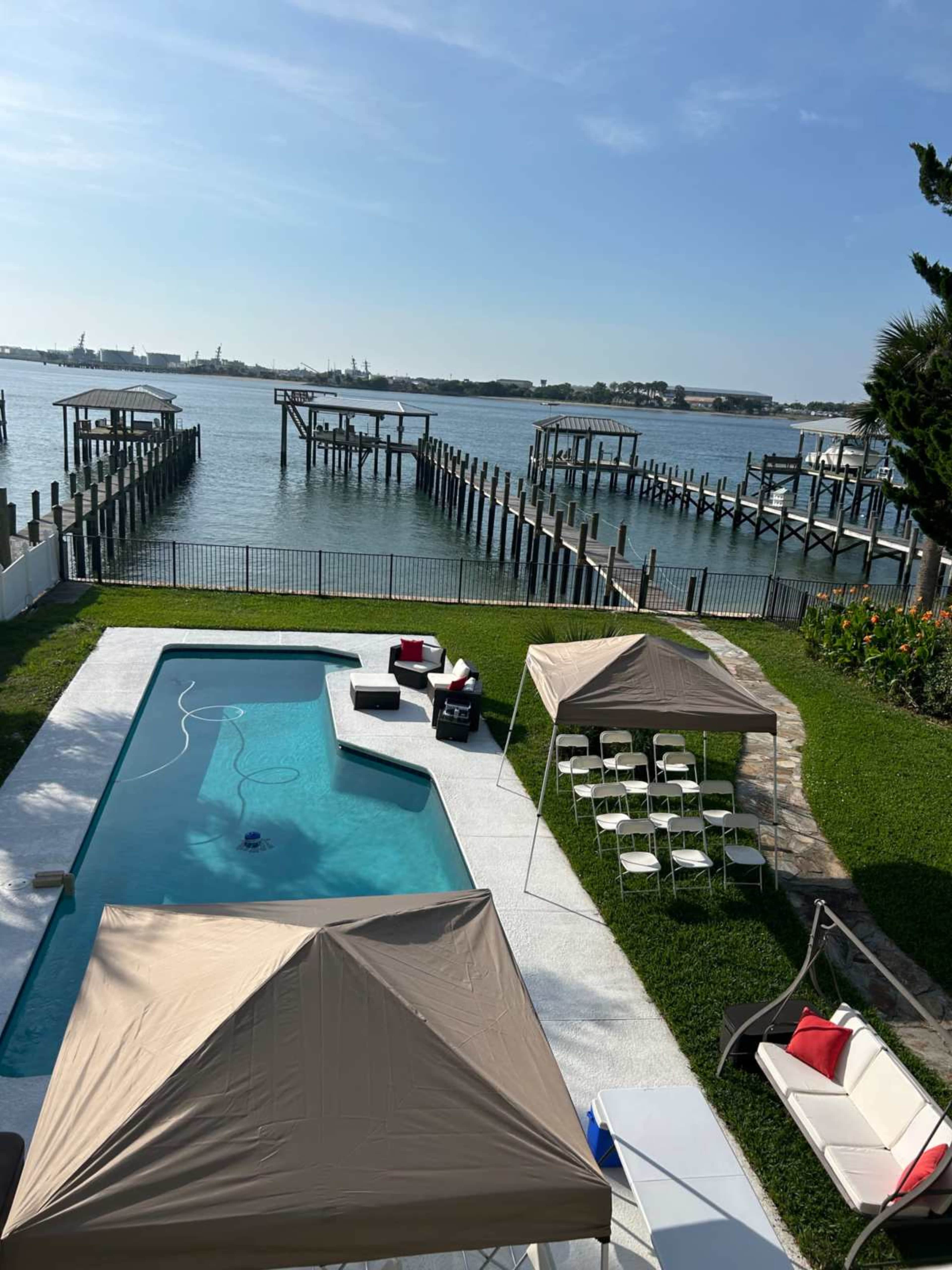The image shows a swimming pool surrounded by grass and tents, with a waterfront view featuring docks and boats in the background.