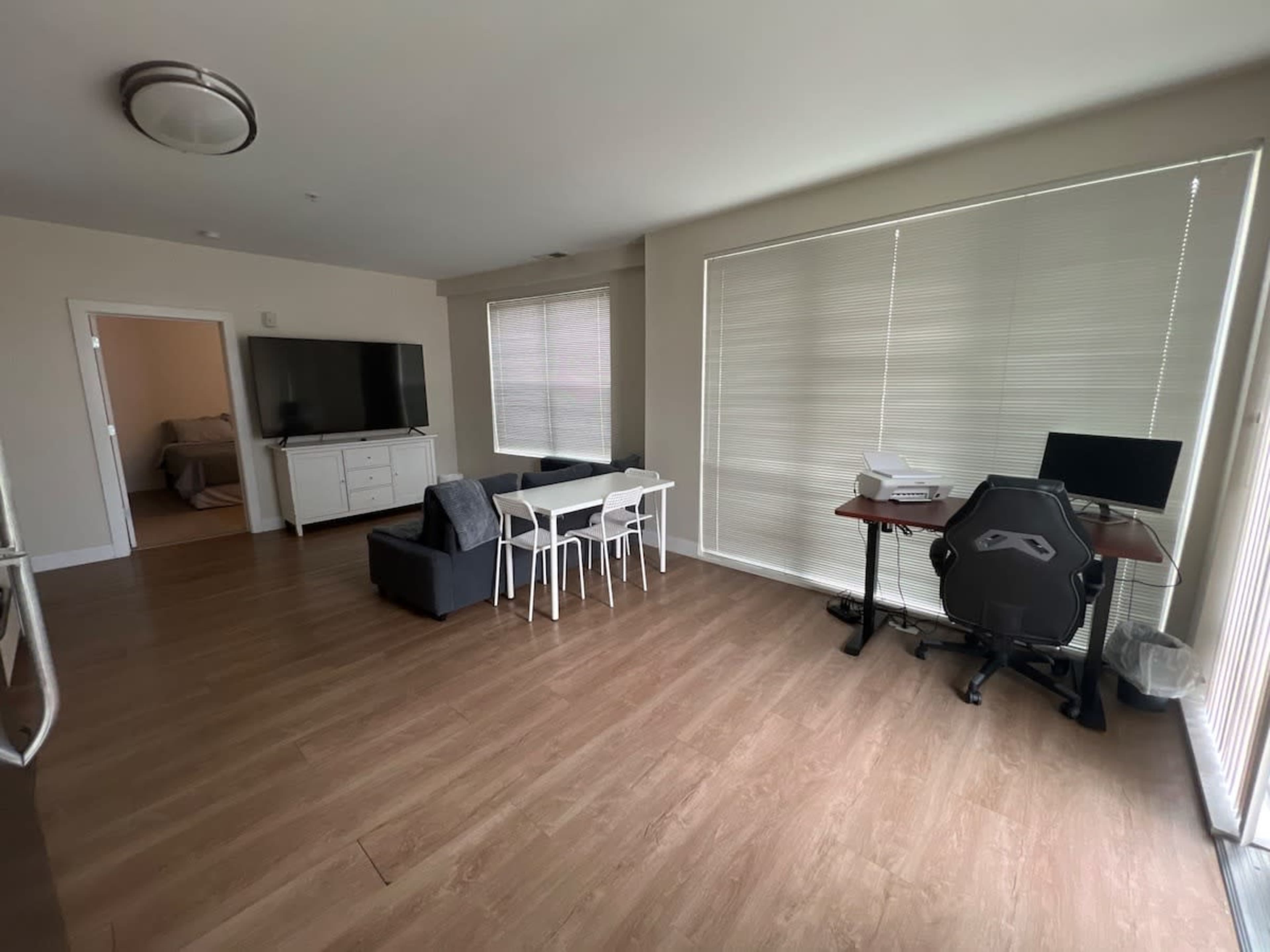 The image shows a modern living room with a television, a small dining table with chairs, a desk with a computer, and large windows covered by blinds.