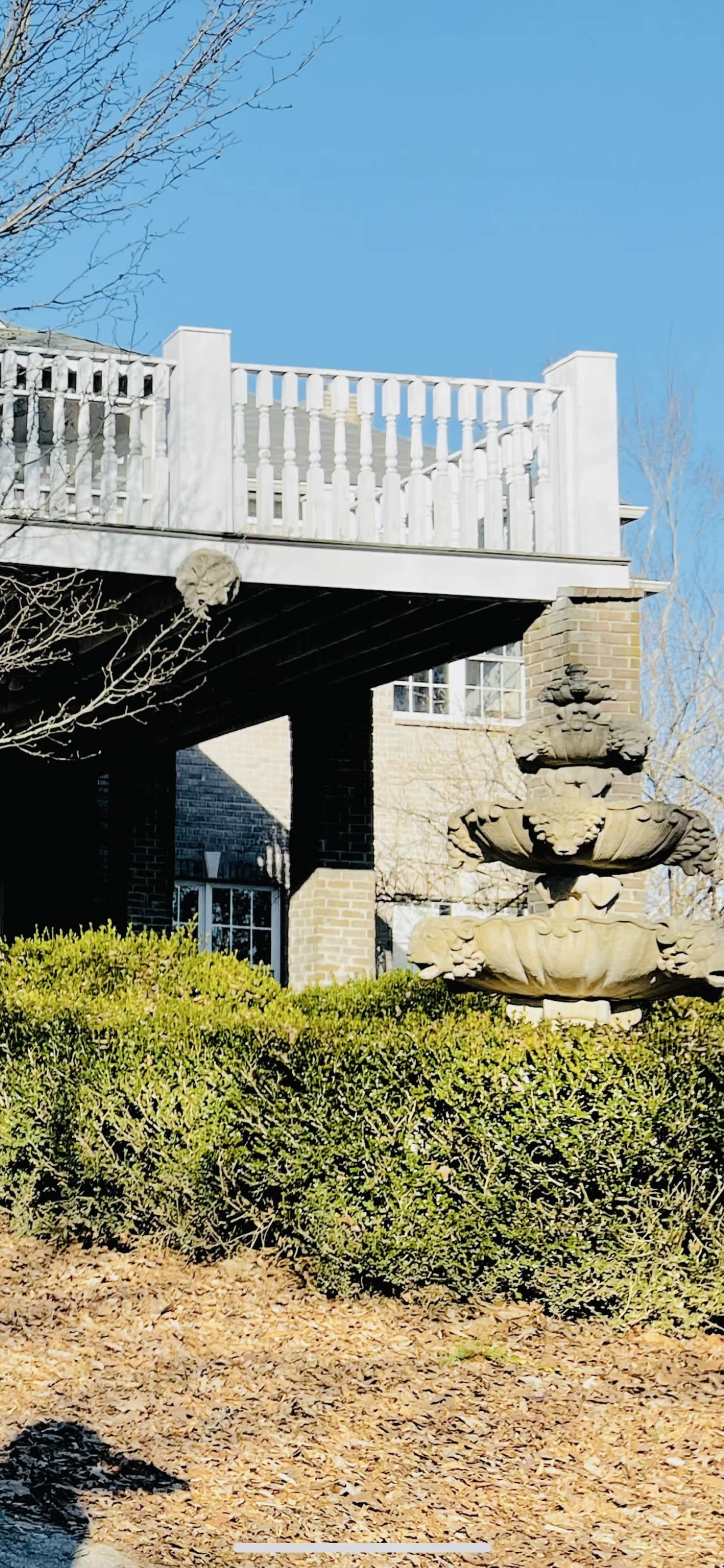 A stone fountain stands in a neatly trimmed garden under a decked balcony of a brick house.