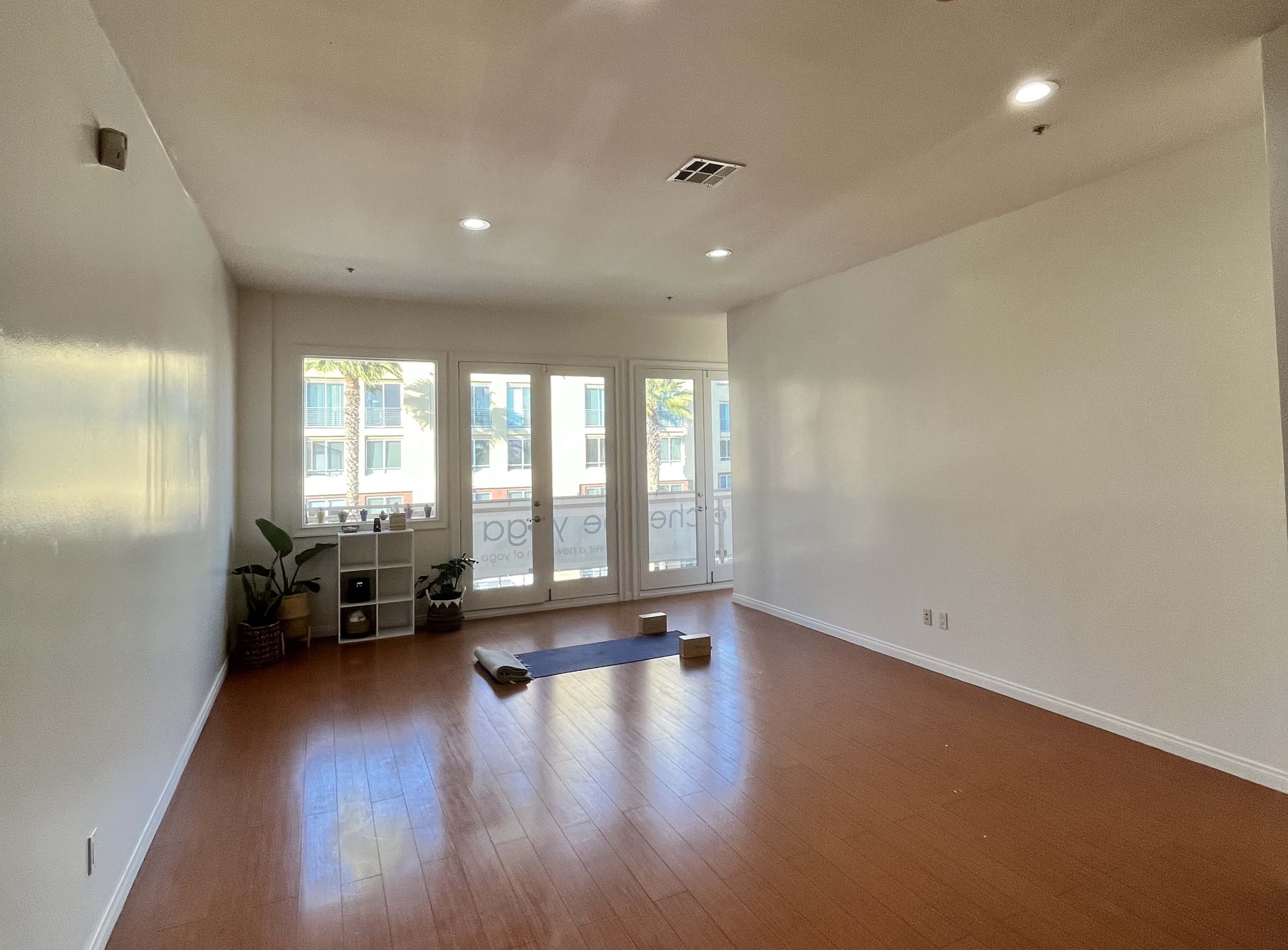 The image shows a spacious, empty room with wooden flooring, a yoga mat, and two yoga blocks, featuring large windows that allow natural light and view of palm trees outside.