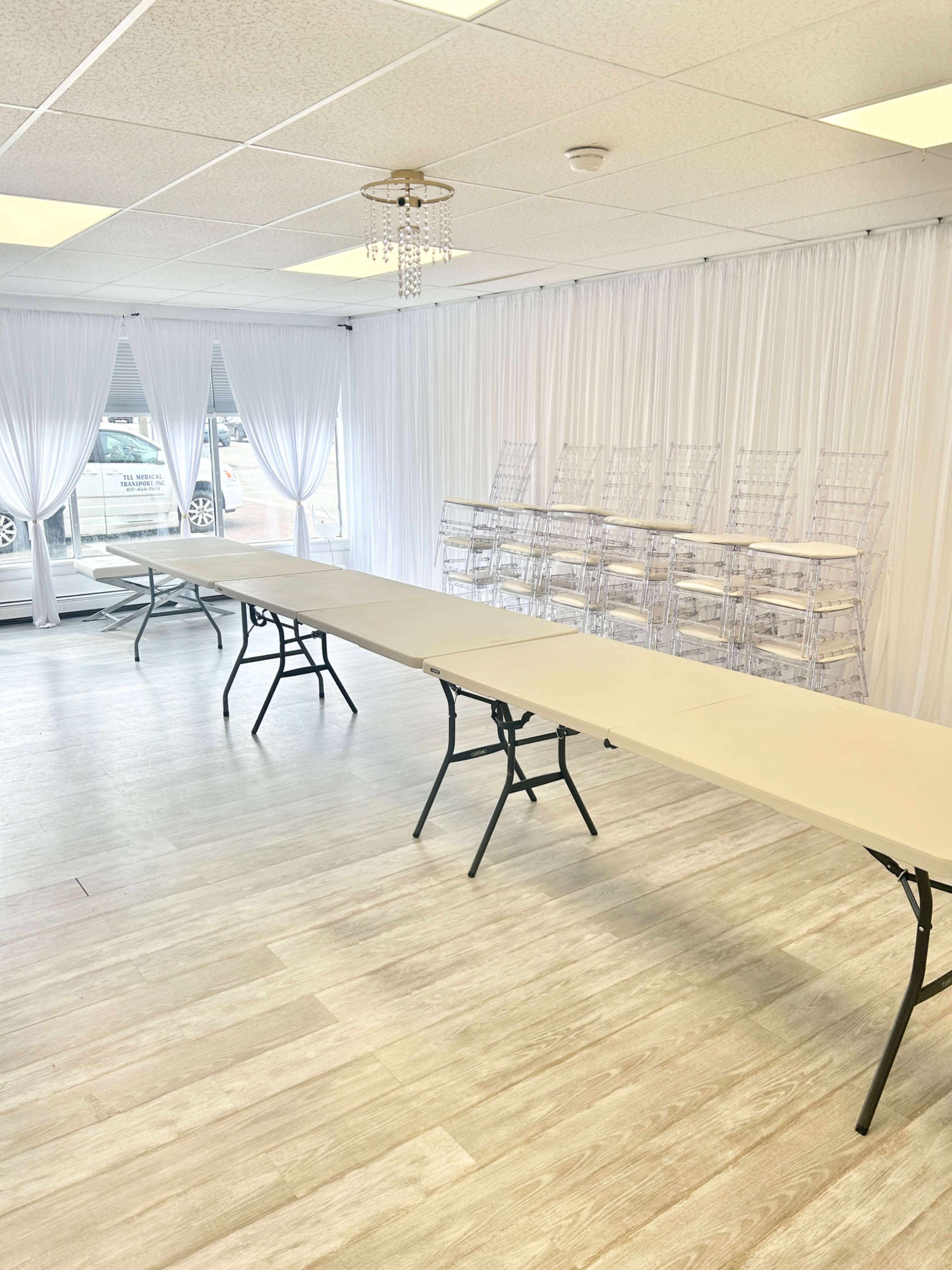 A bright, spacious room features a long folding table set with chairs and sheer white drapes, with several clear acrylic display racks against the wall.