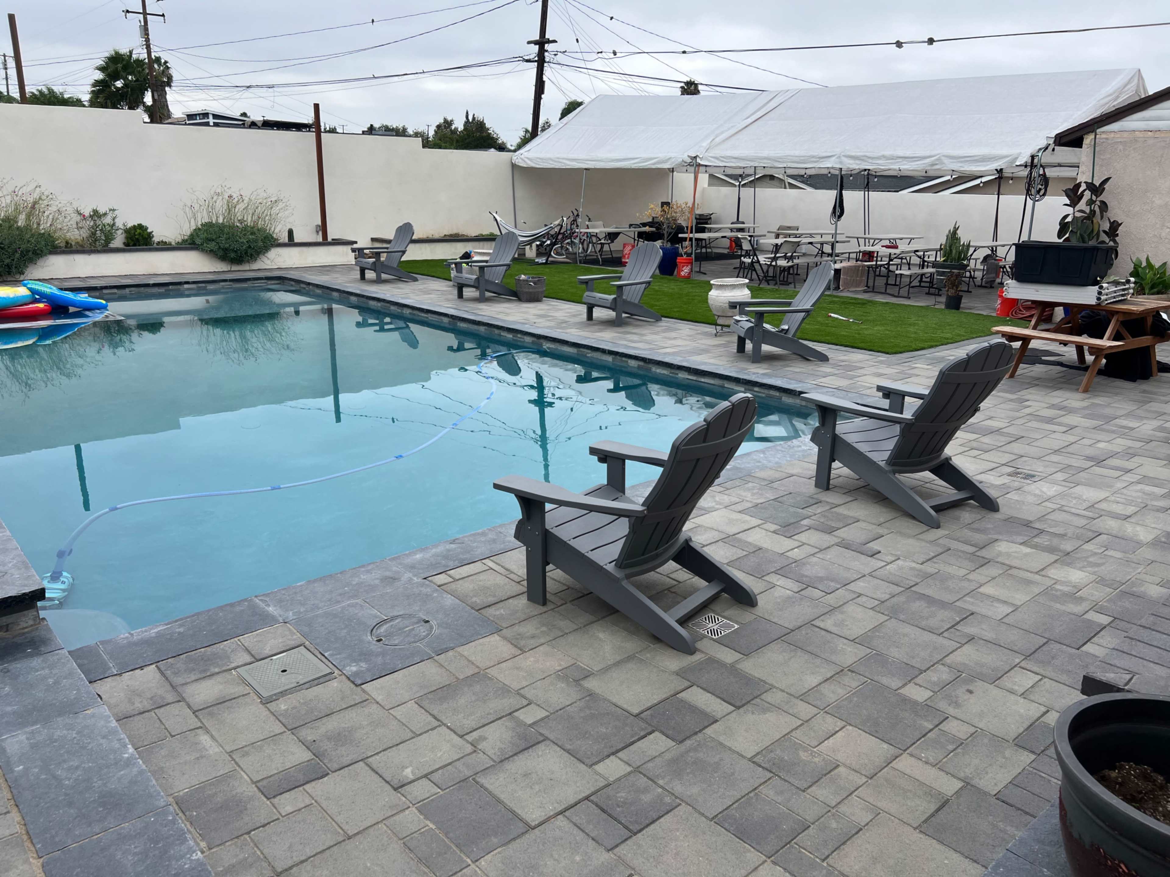 A swimming pool surrounded by gray pavers, with several adirondack chairs positioned nearby and a white tent set up in the background.