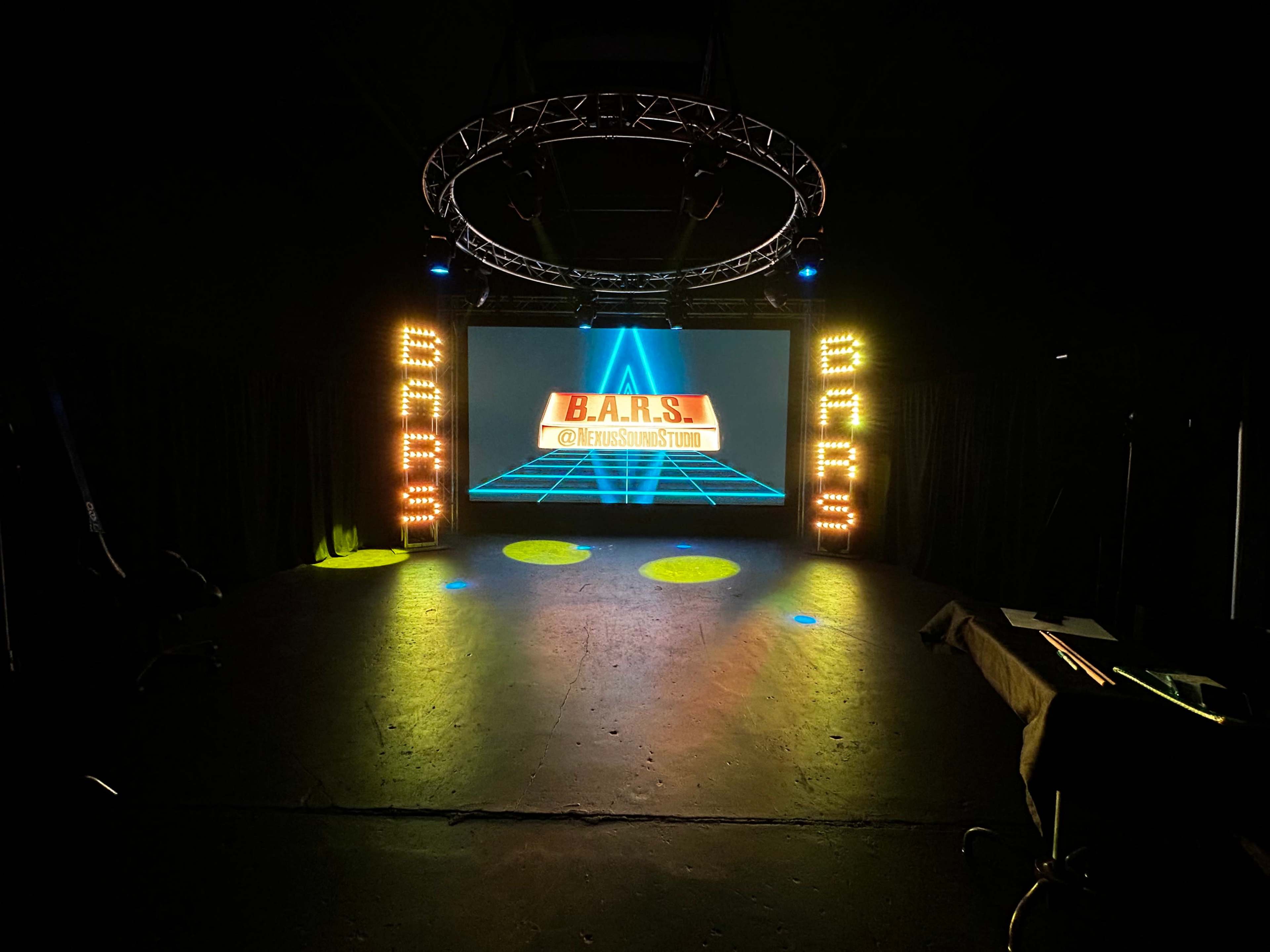 The image shows a stage setup with a large backdrop displaying "B.A.R.S." and colorful lighting on either side.