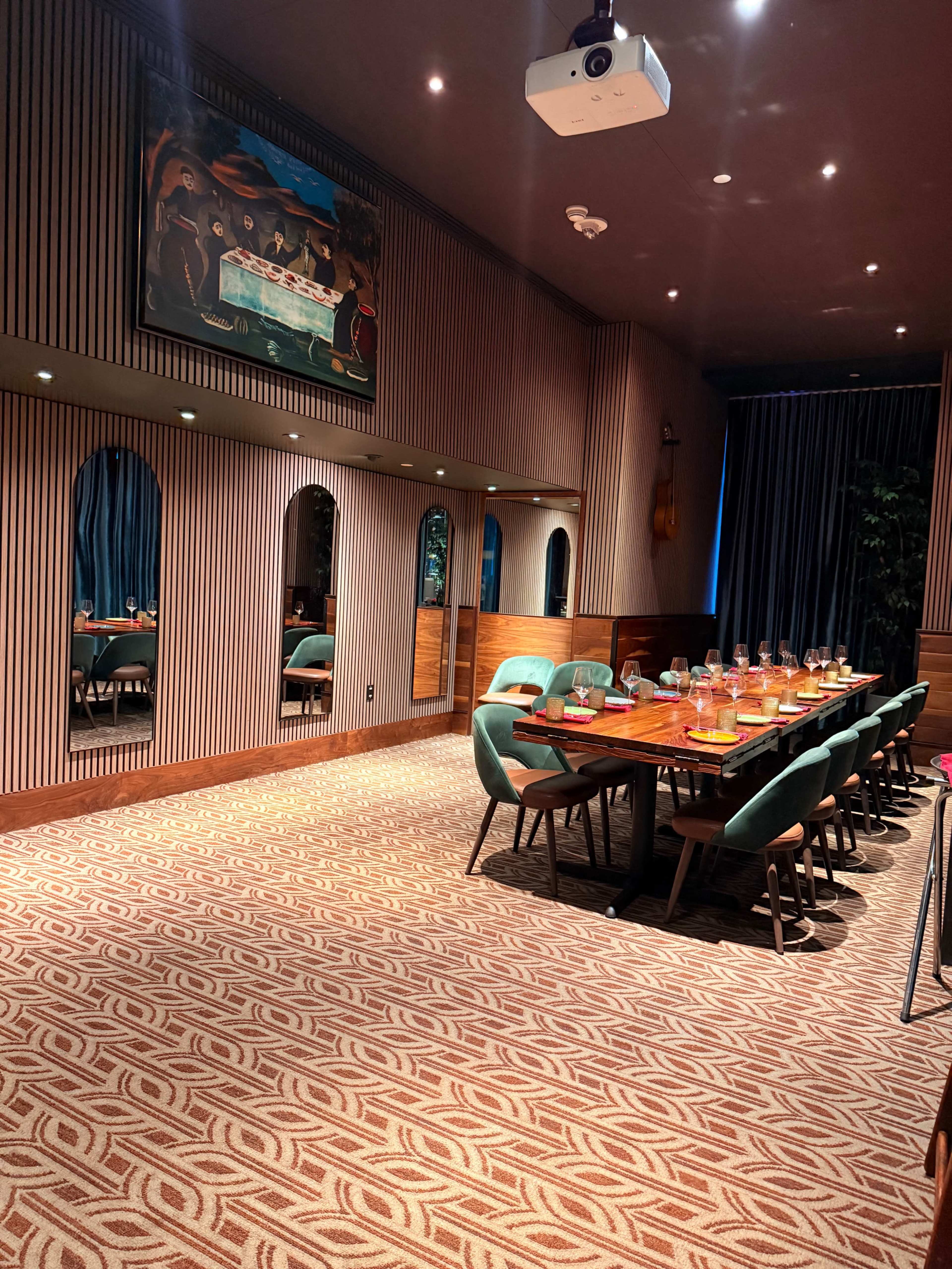 A dining room features a long table with green chairs, illuminated by overhead lighting, and decorated with a large painting and mirrors along the walls.