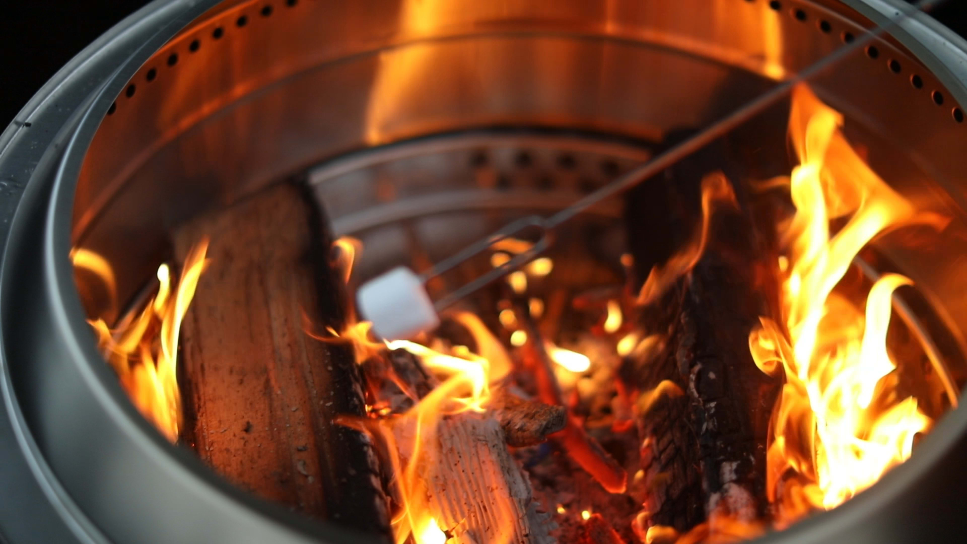 A close-up of a fire with flames engulfing logs and a marshmallow being roasted on a skewer amid the glowing embers.