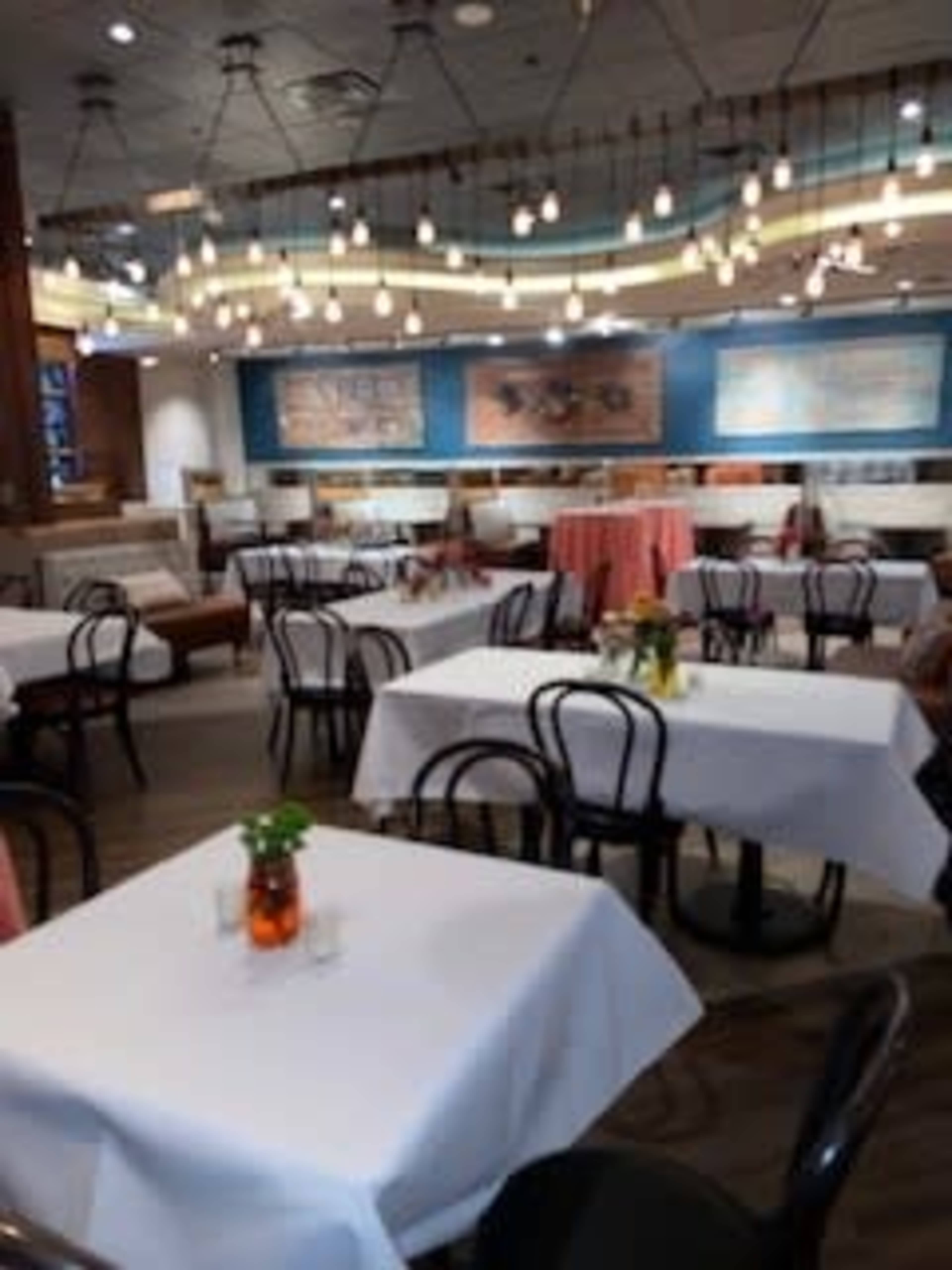 The image shows a restaurant interior featuring several empty tables with white tablecloths, surrounded by decorative light fixtures and vibrant wall art.