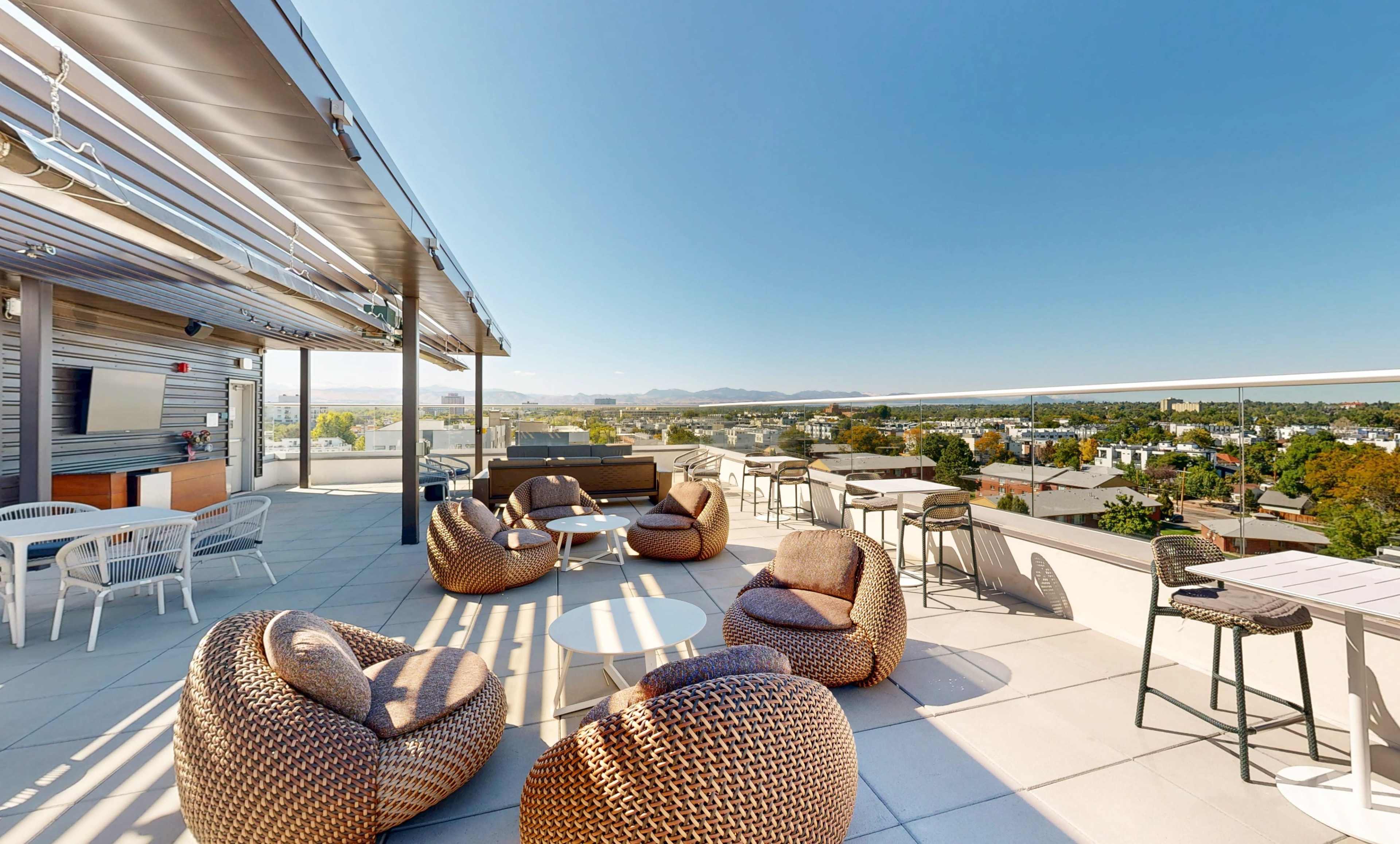 A rooftop terrace features various seating options, including round tables and woven lounge chairs, overlooking a panoramic view of the surrounding landscape.