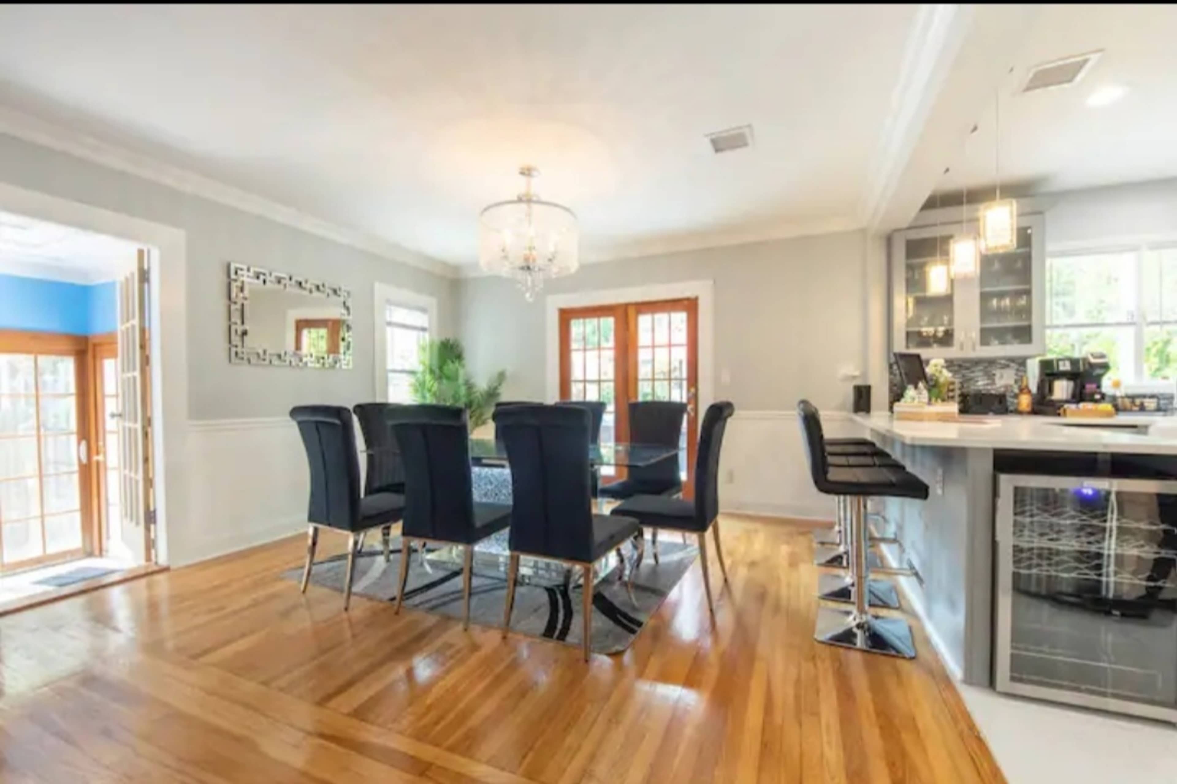 The image shows a modern dining area with a large table surrounded by black chairs, adjacent to a well-equipped kitchen and featuring hardwood floors.