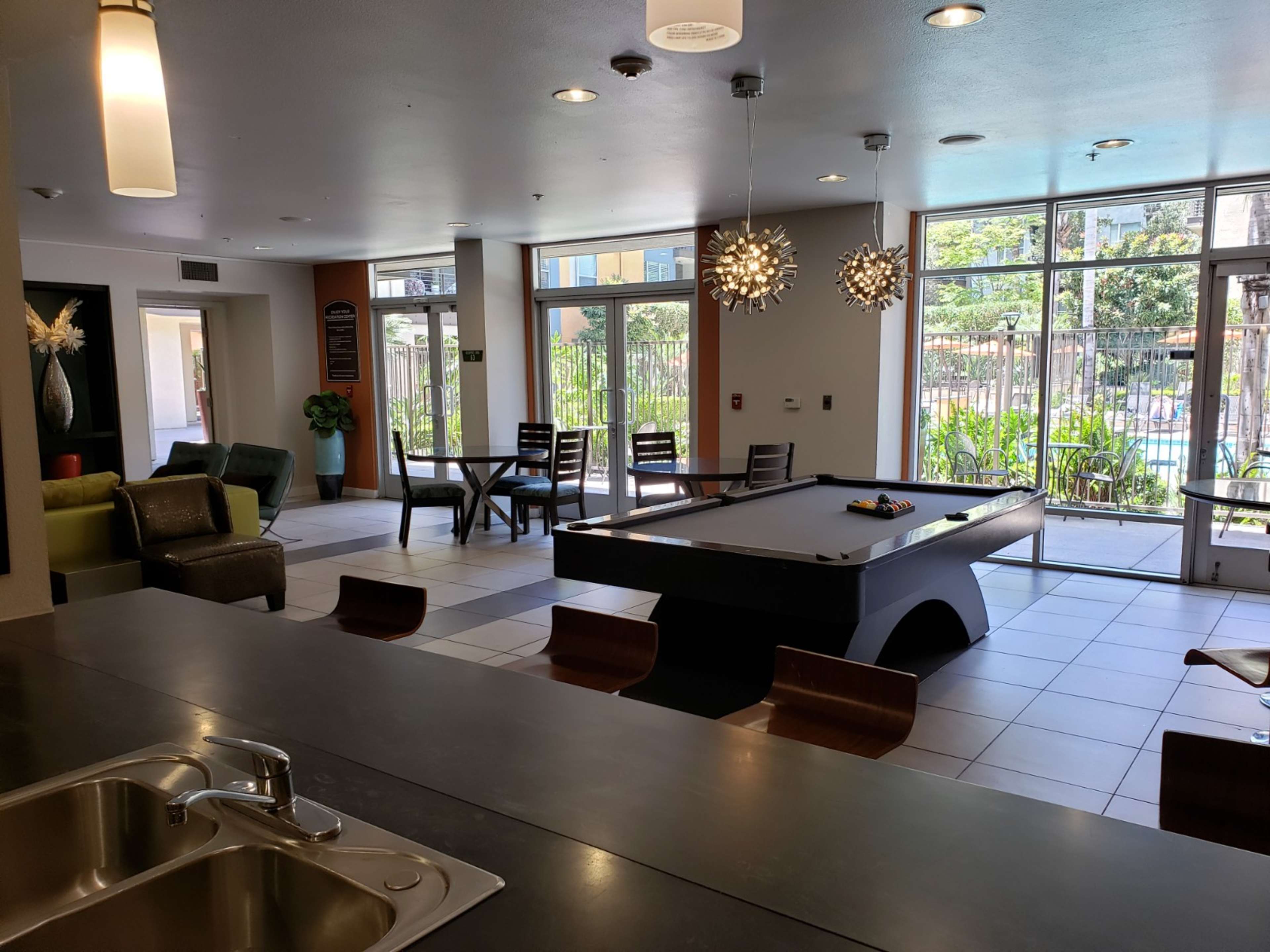 A common area with a pool table, dining tables, and large windows overlooking a pool and outdoor seating.
