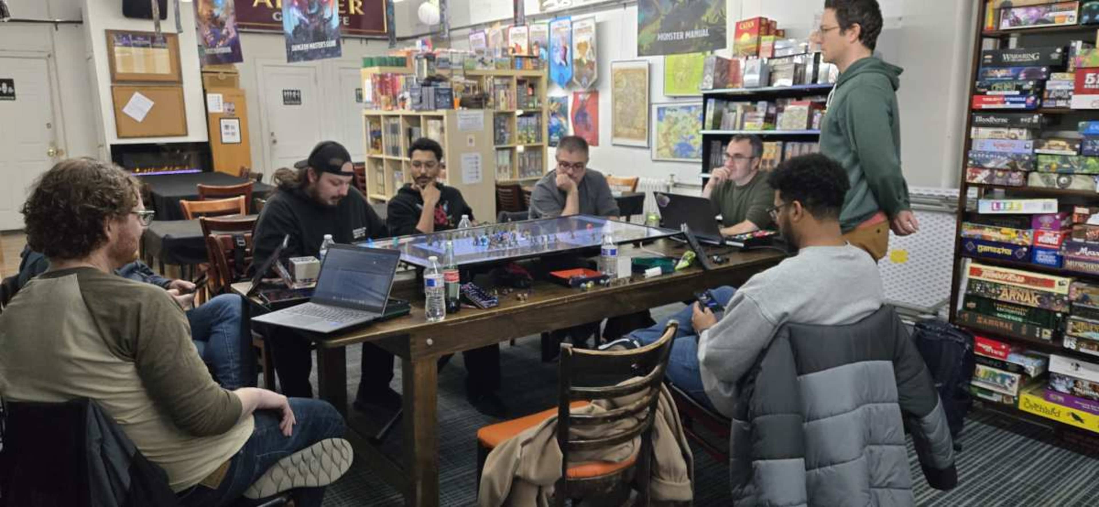 A group of people sits around a large table in a room filled with shelves of board games, engaged in a gaming session with laptops open in front of them.