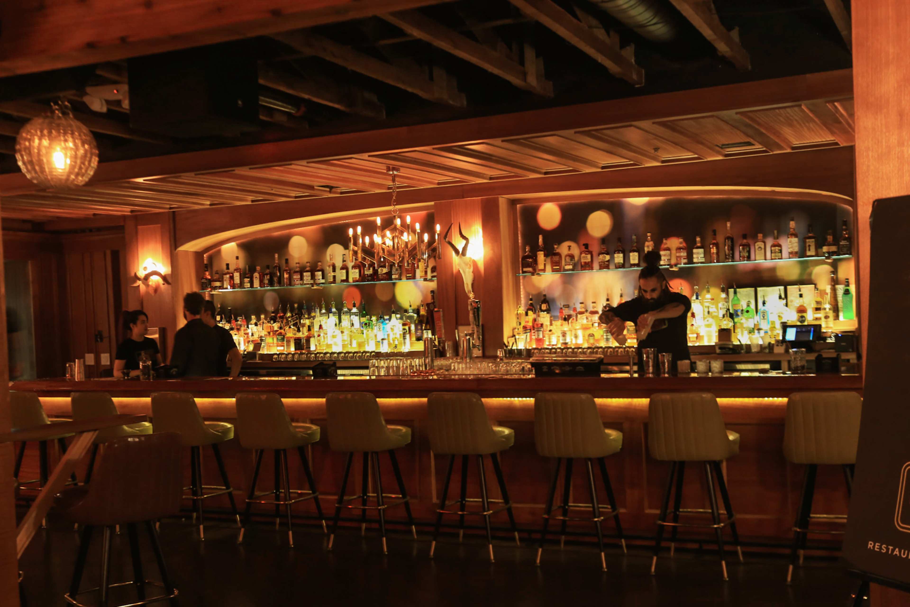 A dimly lit bar features a well-stocked back shelf with bottles and two bartenders preparing drinks.