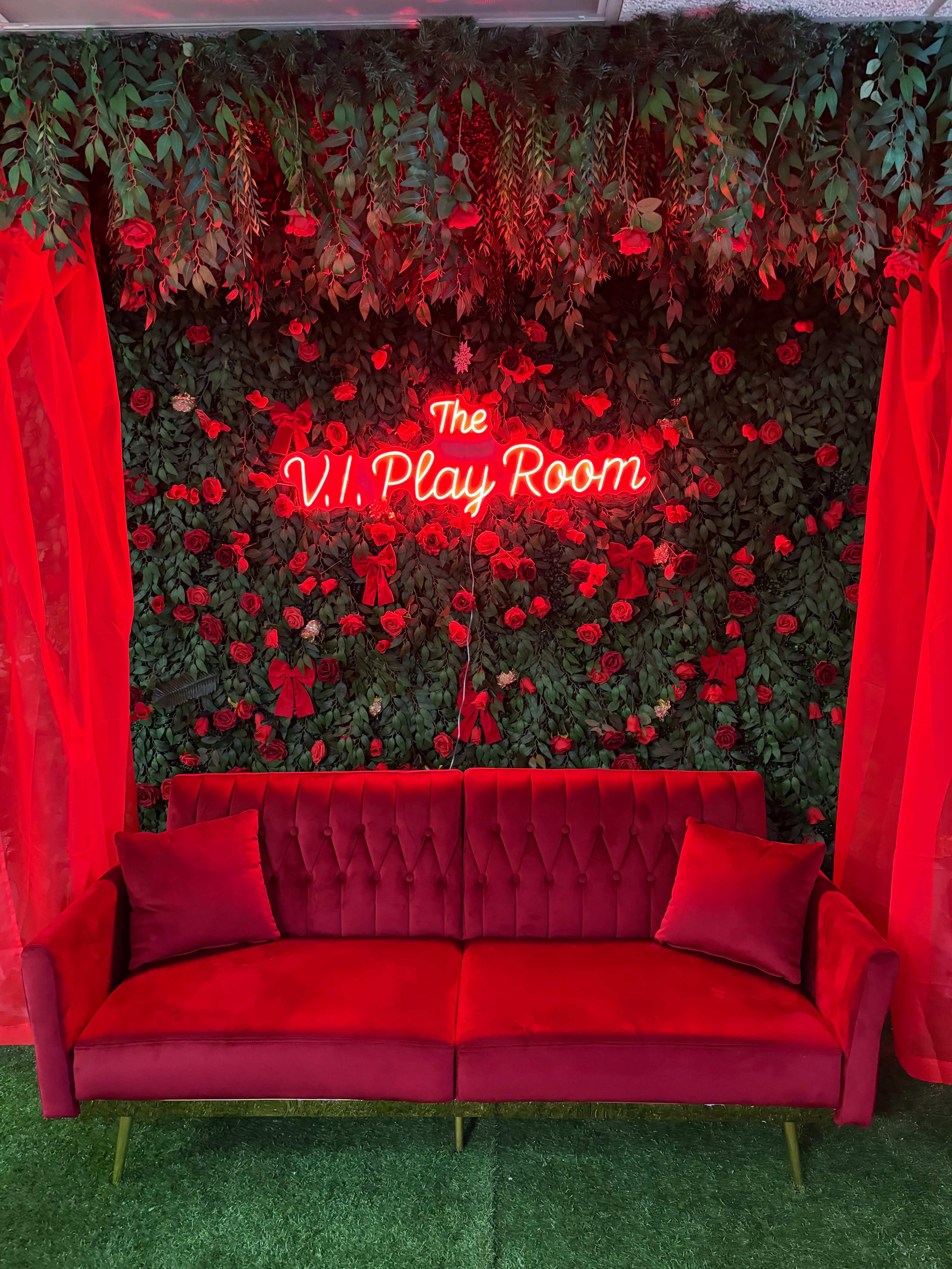 A red velvet sofa is positioned in front of a floral wall adorned with red roses and greenery, featuring a neon sign that reads "The V.I. Play Room."