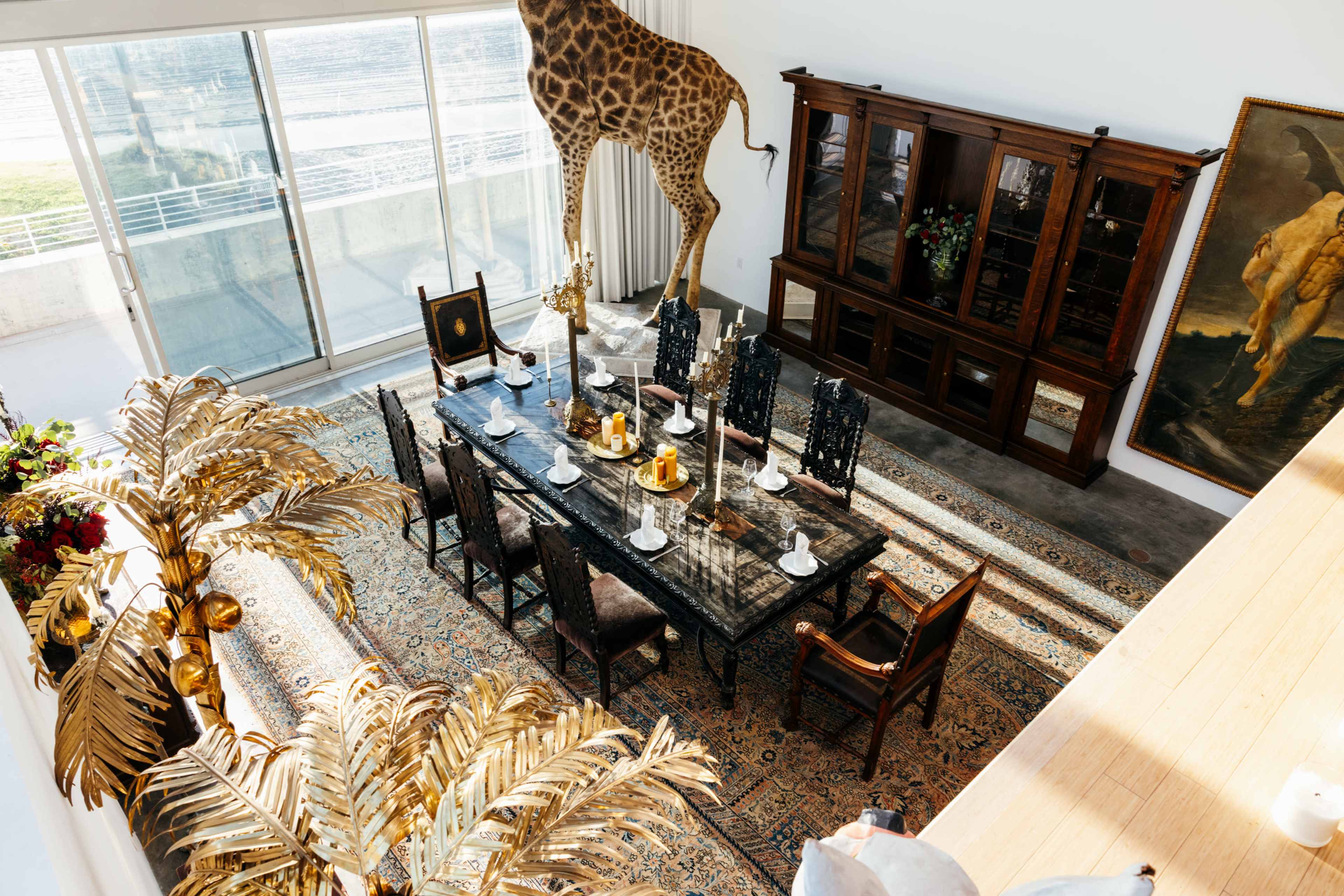 The image depicts an elegantly set dining room with a long table surrounded by chairs, featuring a giraffe sculpture and large windows allowing natural light to fill the space.