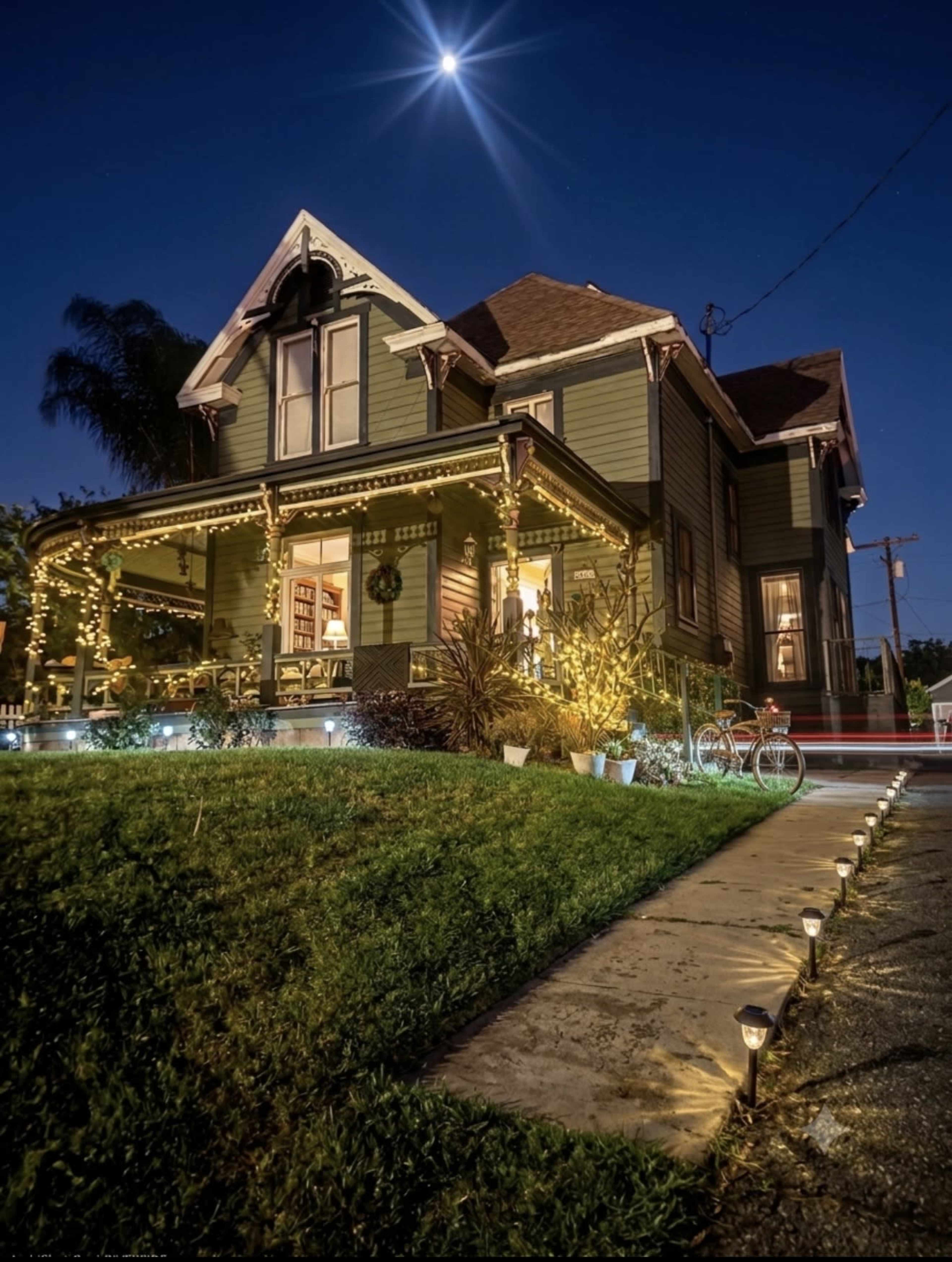 A well-lit, two-story Victorian-style house is adorned with string lights and sits under a night sky featuring a bright moon.