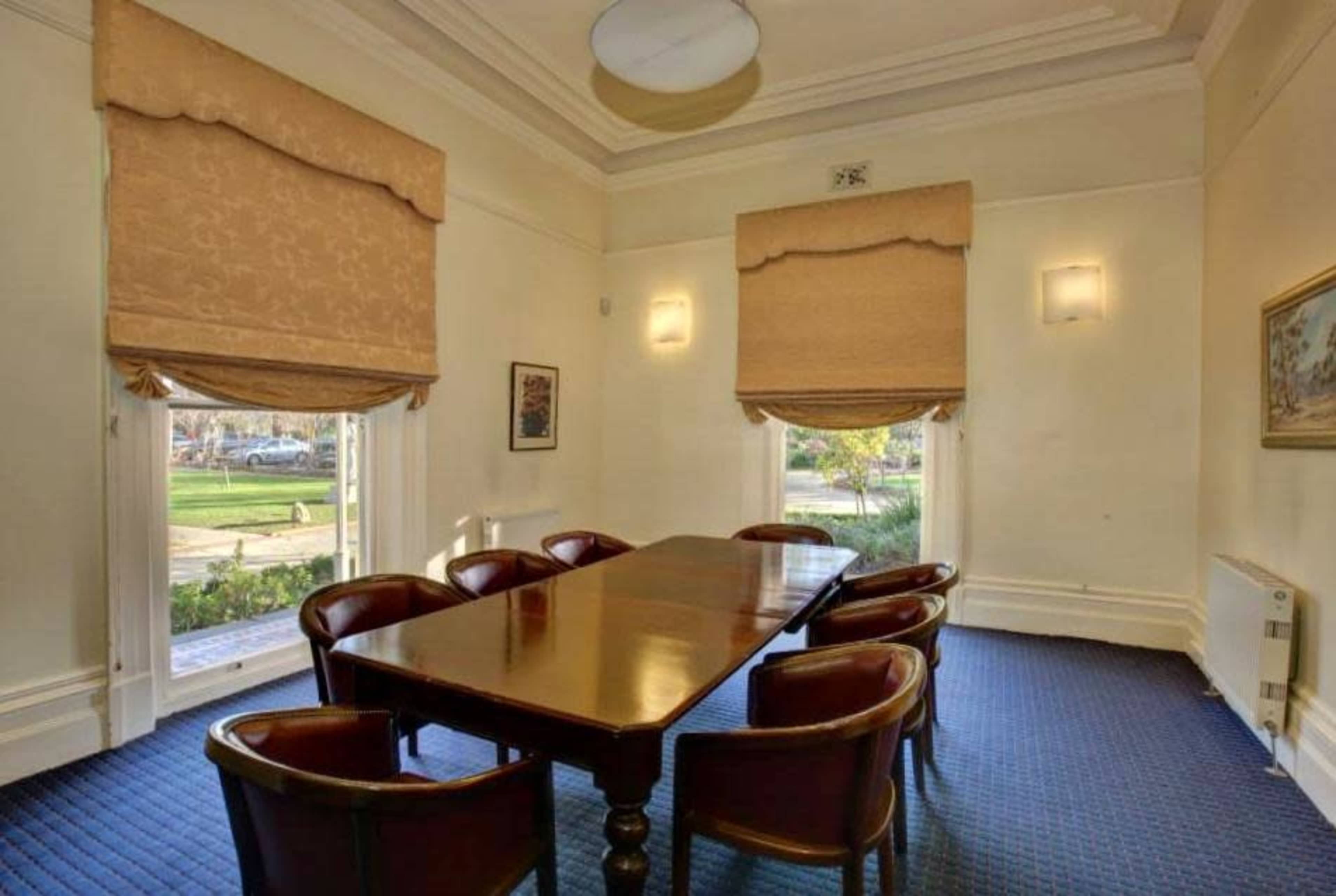 A well-lit meeting room features a long wooden table surrounded by six chairs, with large windows covered by light-colored shades.