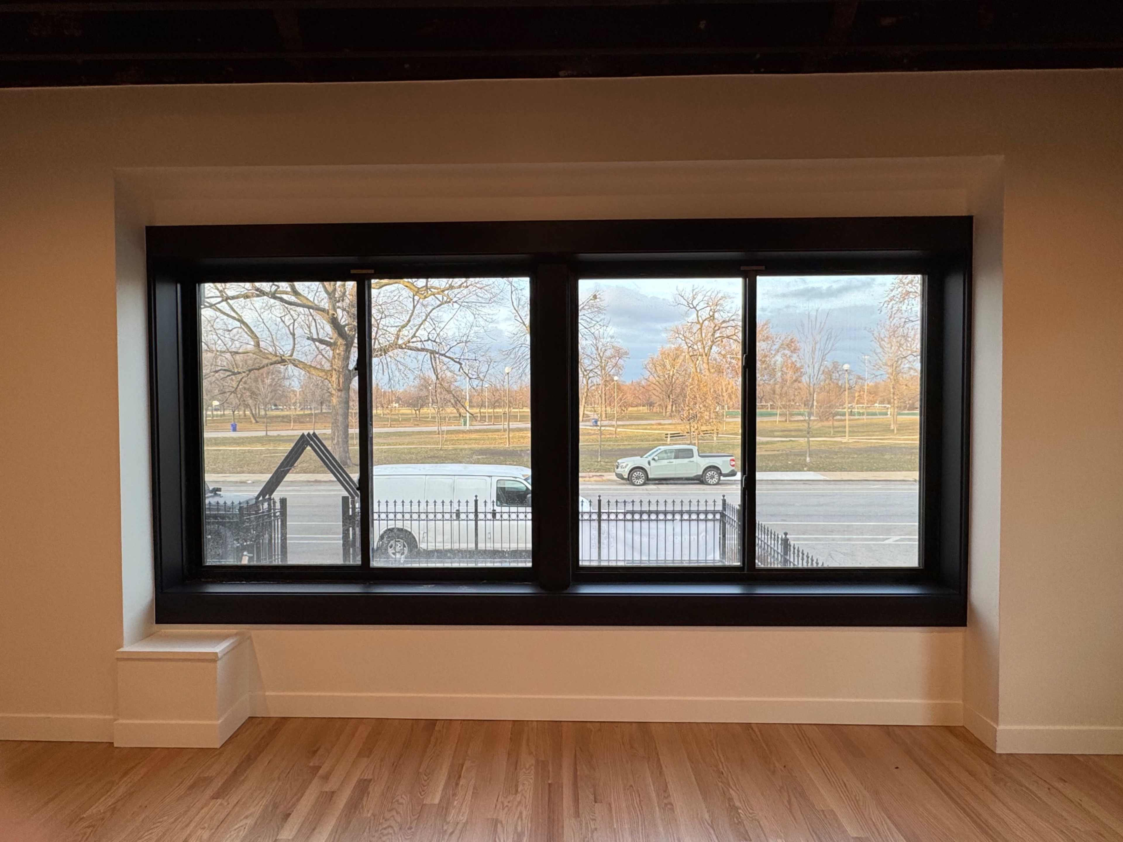The image shows a large, black-framed window with three panes, overlooking a road with trees and parked vehicles outside.