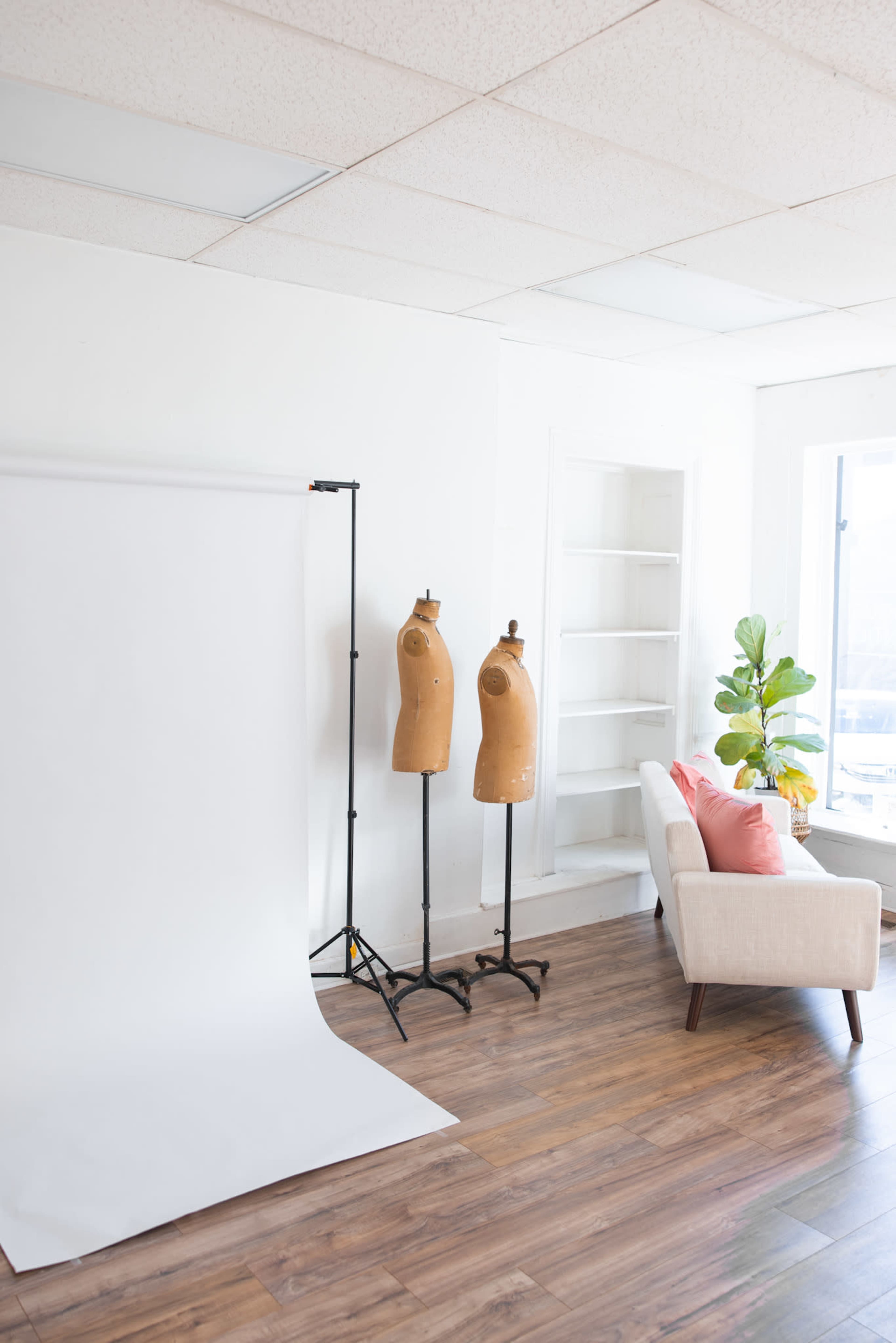 The image features a bright, minimalist room with a white backdrop stand, two wooden dress forms, and a light-colored sofa adorned with a pink cushion.