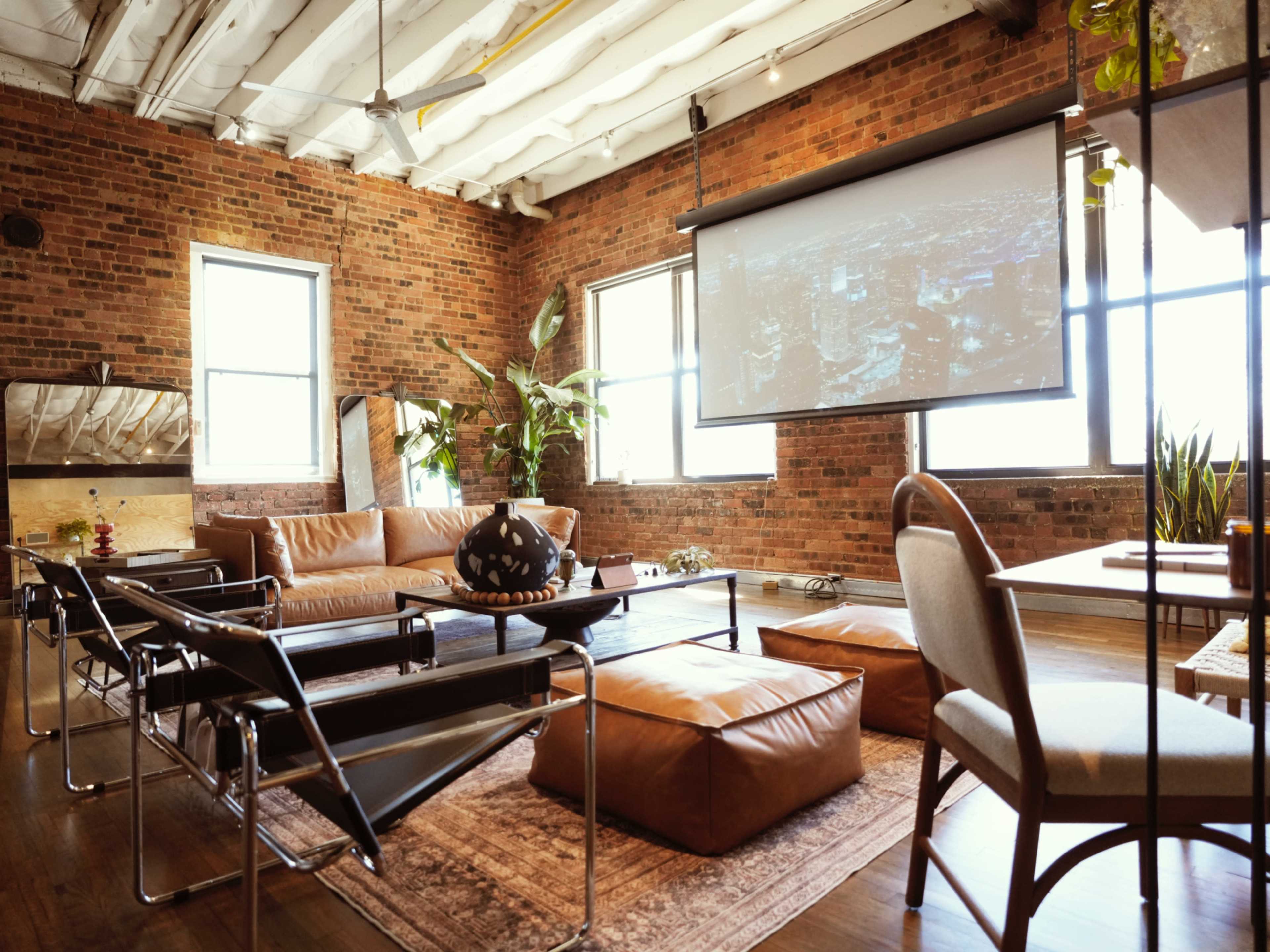 The scene shows a modern living room with exposed brick walls, a leather sofa, and a projector screen displaying an image, complemented by plants and various seating options.