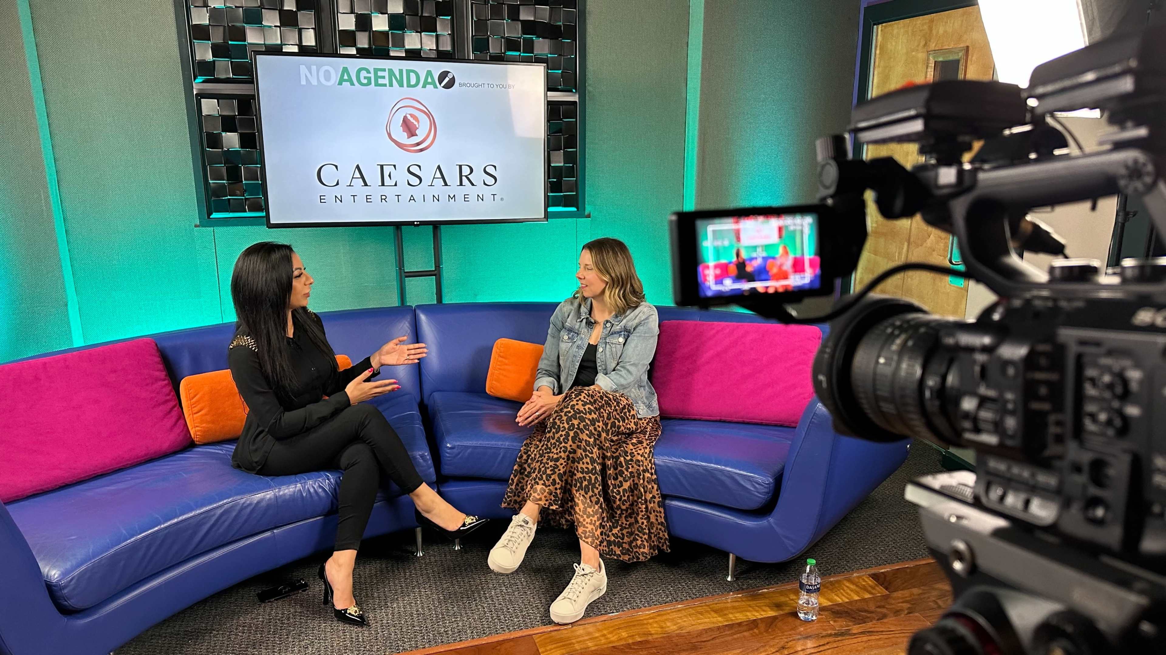 Two women sit on a colorful couch in a studio, engaged in conversation, with a camera and a logo for Caesars Entertainment visible in the background.