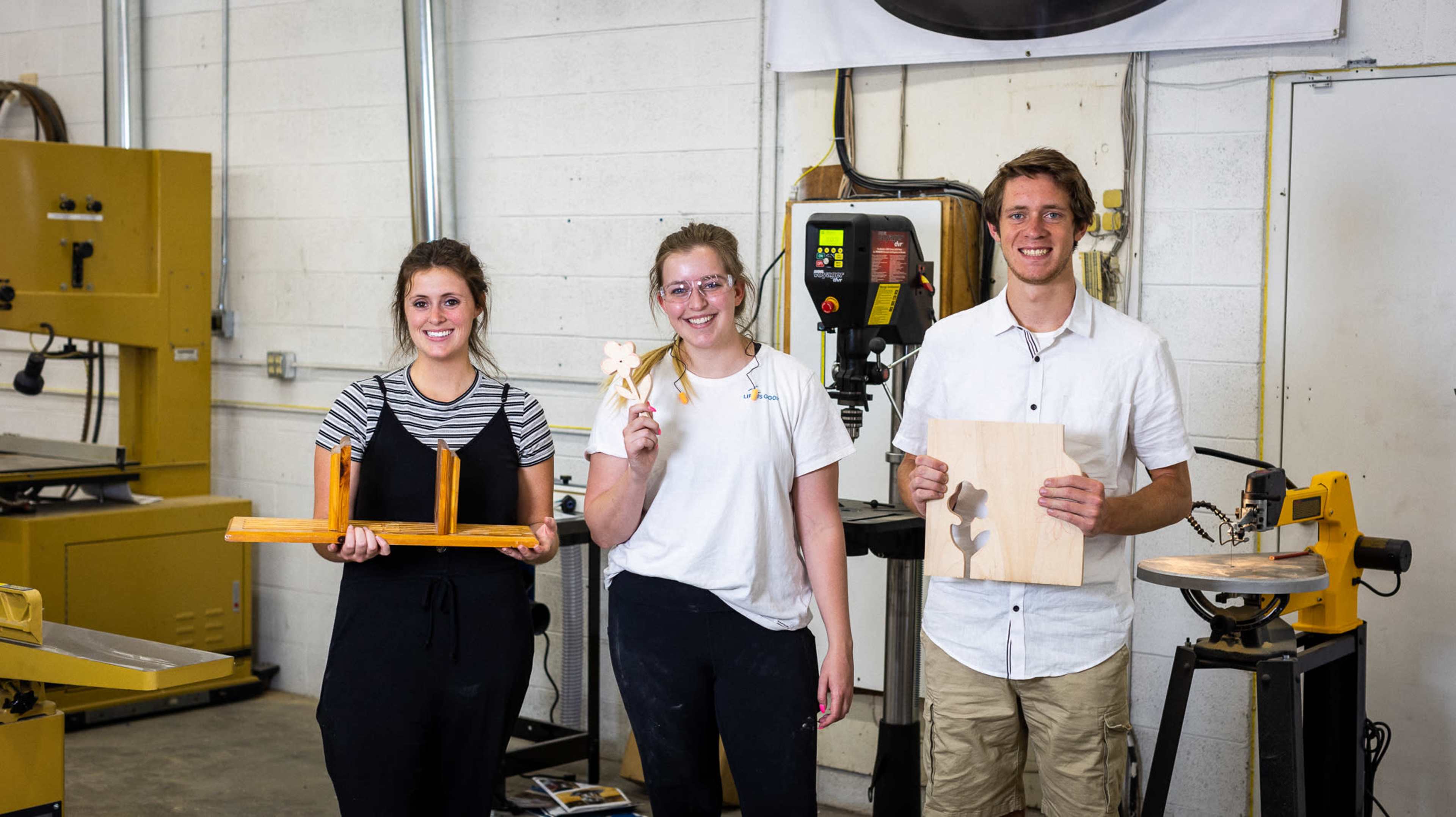 Three individuals stand in a workshop holding wooden projects, with various woodworking machines in the background.