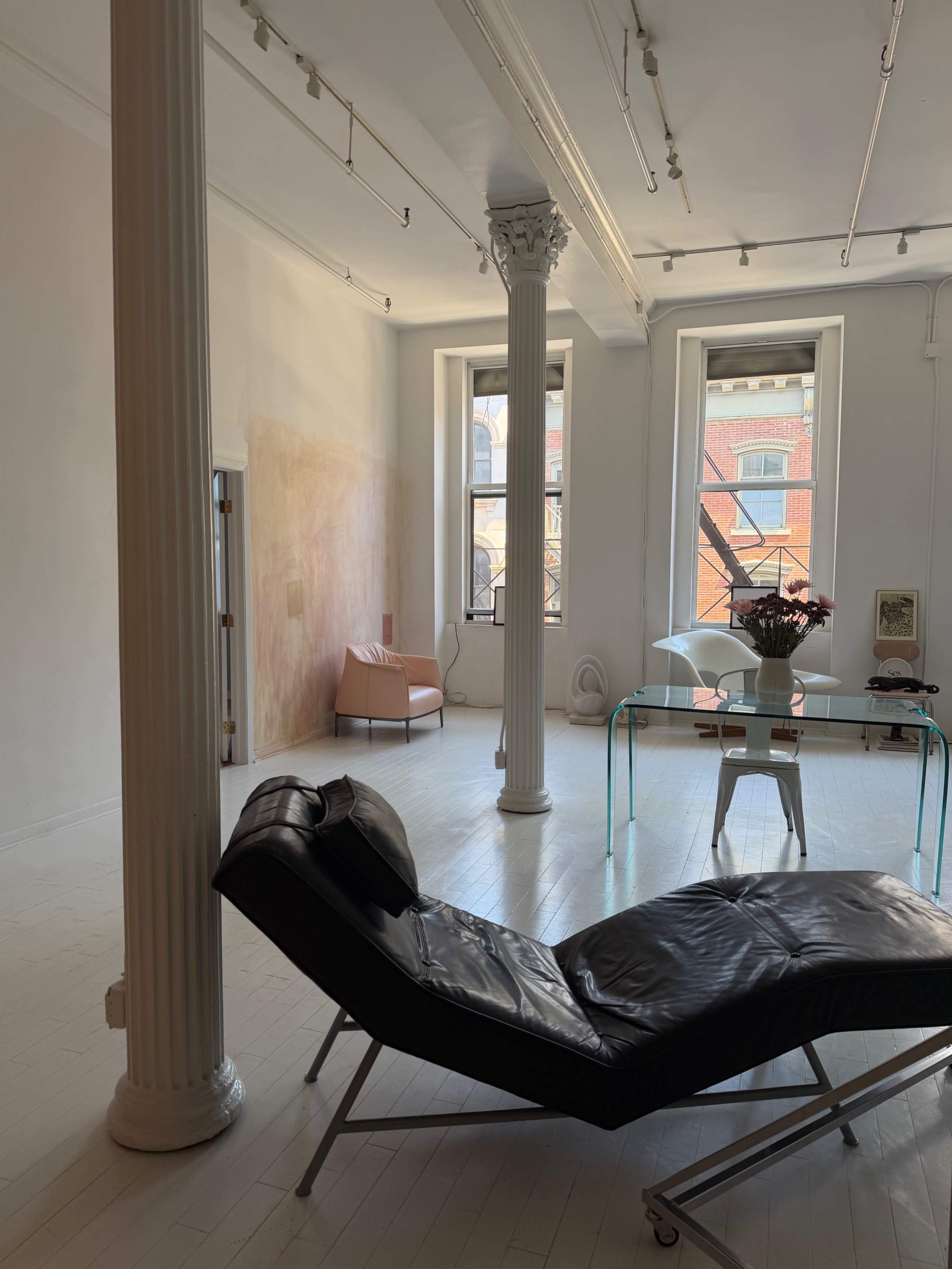 The image shows an airy, minimalist interior with a black chaise lounge, a glass table, and large windows allowing natural light to enter the space.