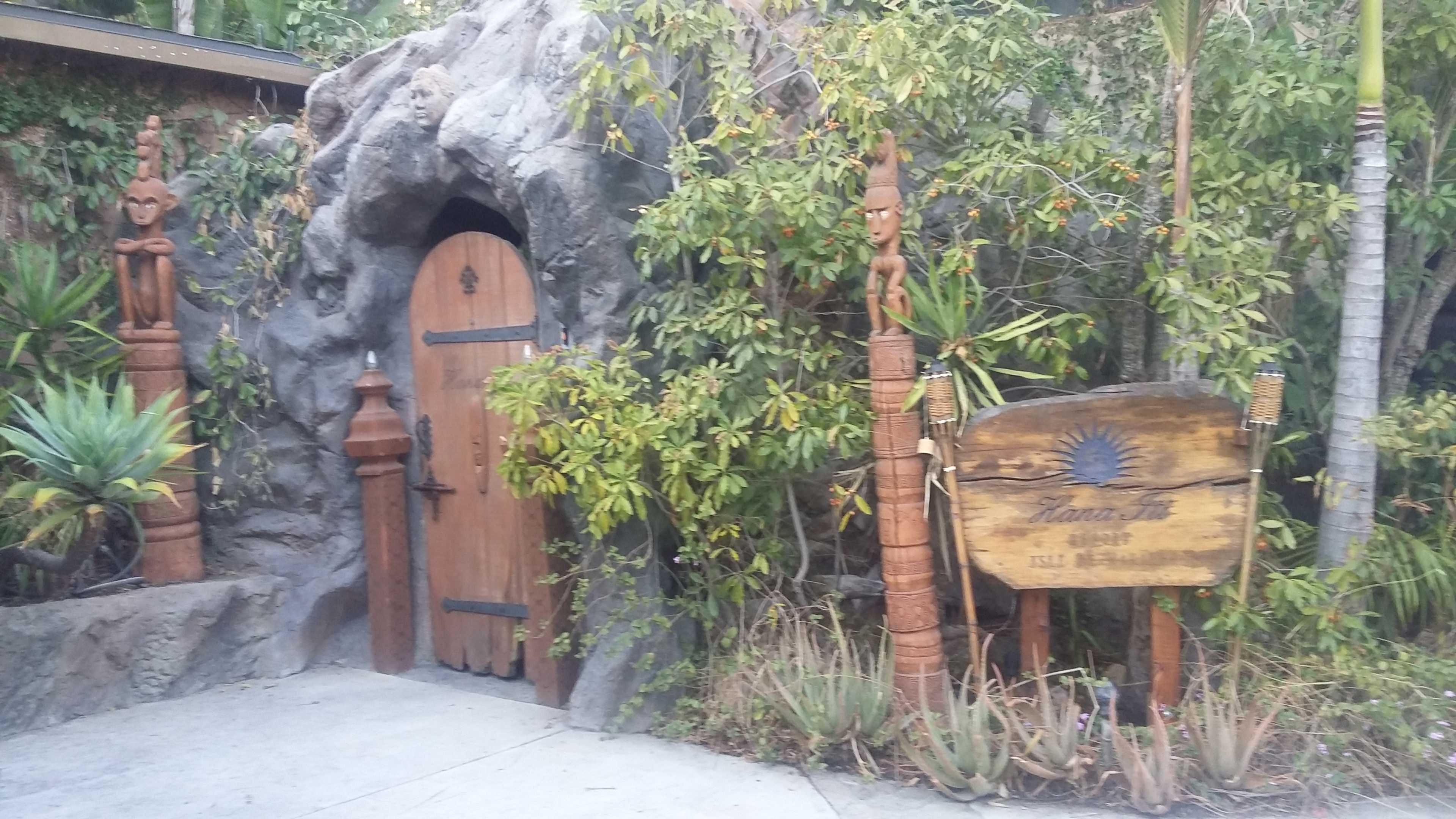 A wooden door set into a rocky entrance is flanked by carved totem poles and surrounded by lush vegetation.
