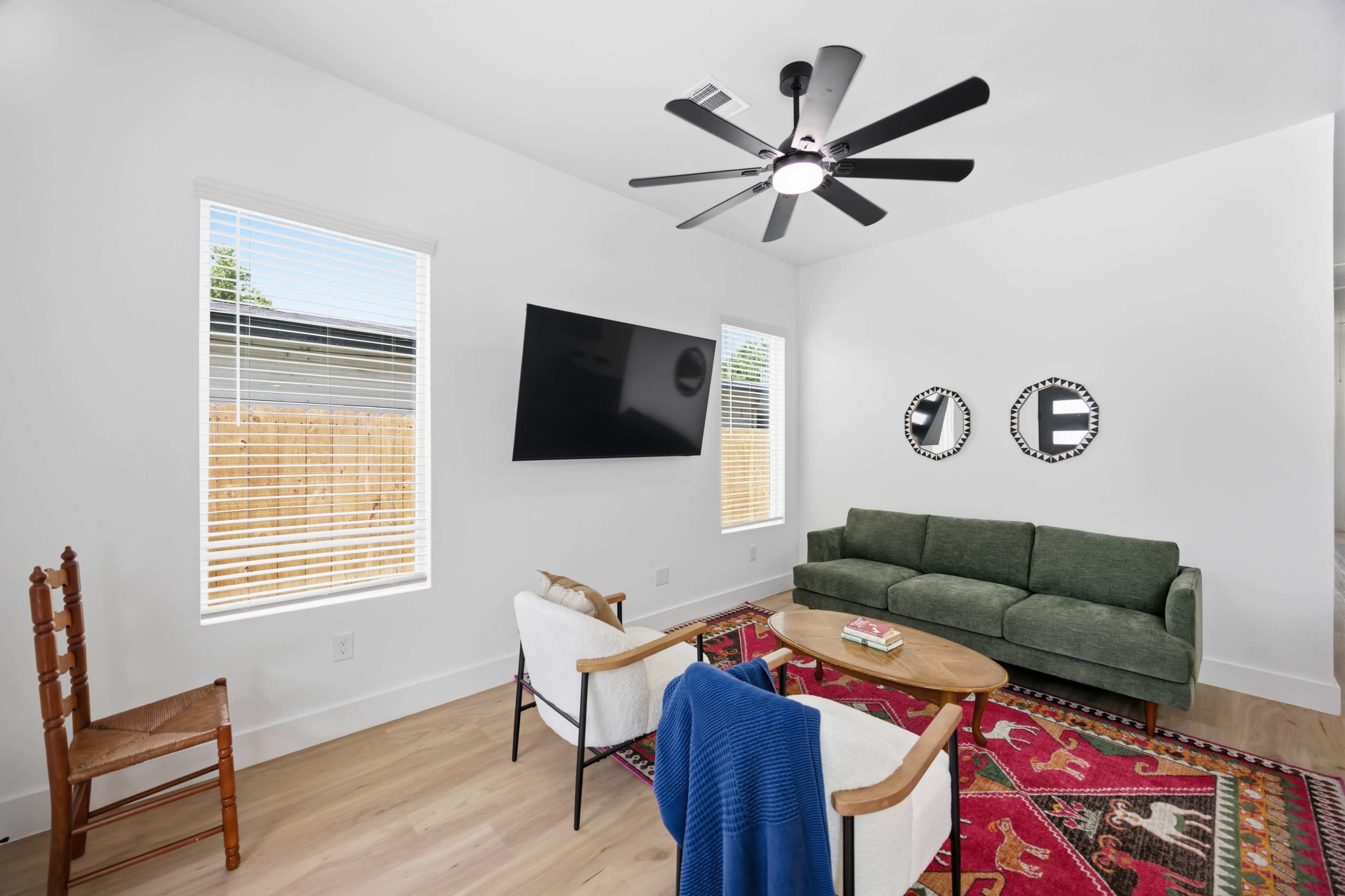 The scene shows a modern living room featuring a green sofa, two upholstered chairs, a round coffee table, a mounted television, and large windows that let in natural light.