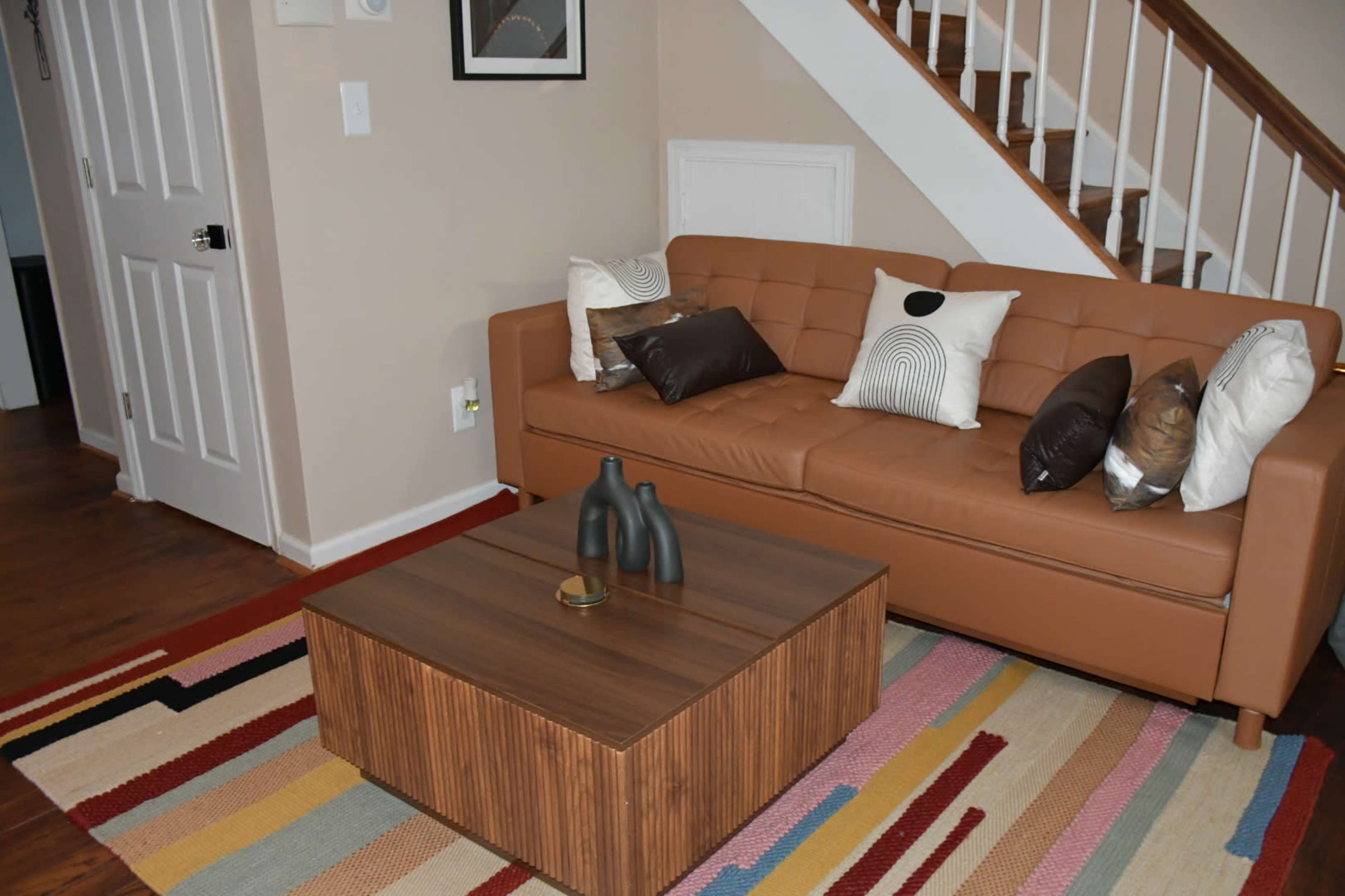 A modern living space features a brown leather sofa with decorative pillows, a wooden coffee table, and a colorful patterned rug, all positioned near a staircase.