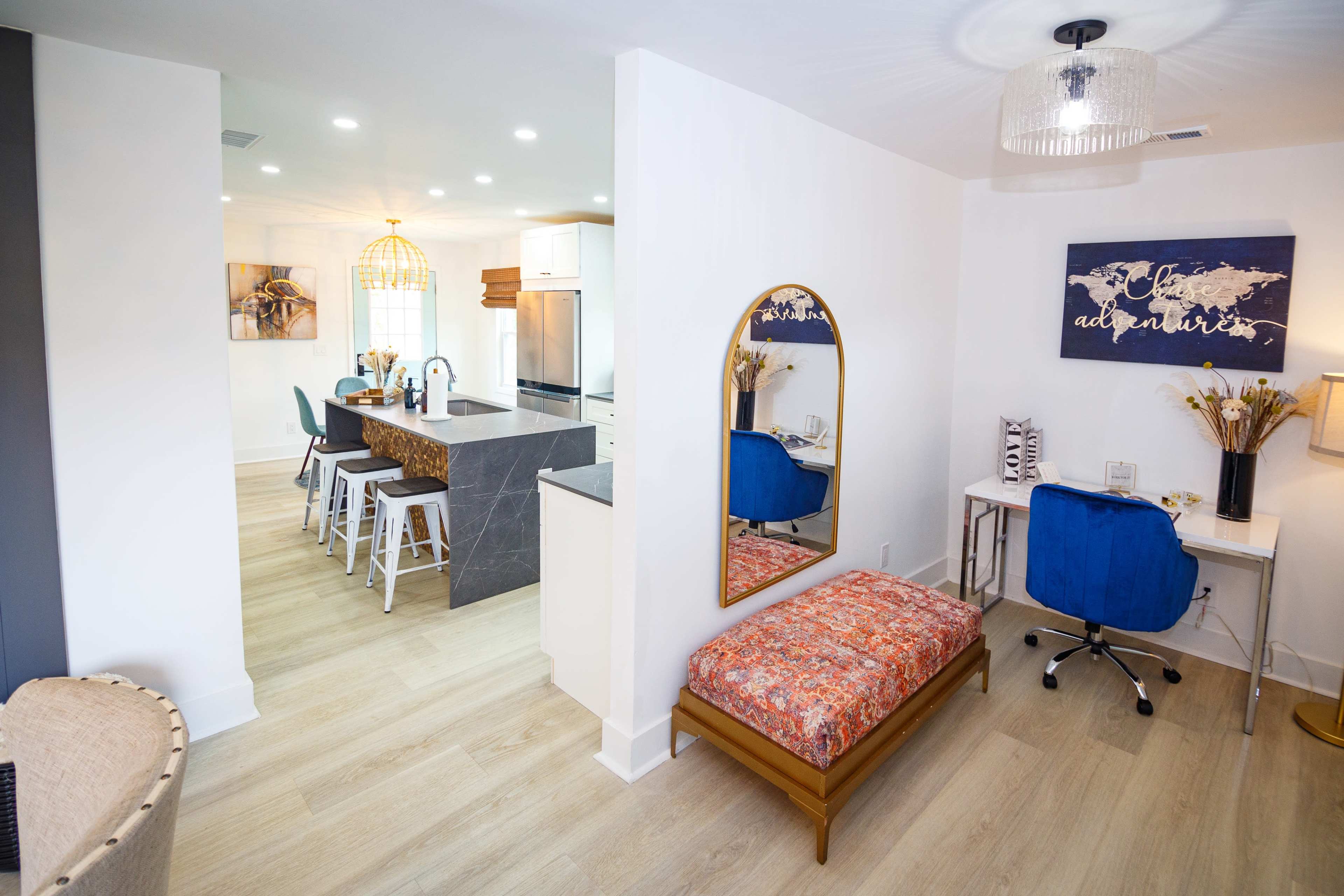 The image shows a bright, modern interior featuring a kitchen with a large island, a small workspace with a desk and chair, and a decorative bench against the wall.