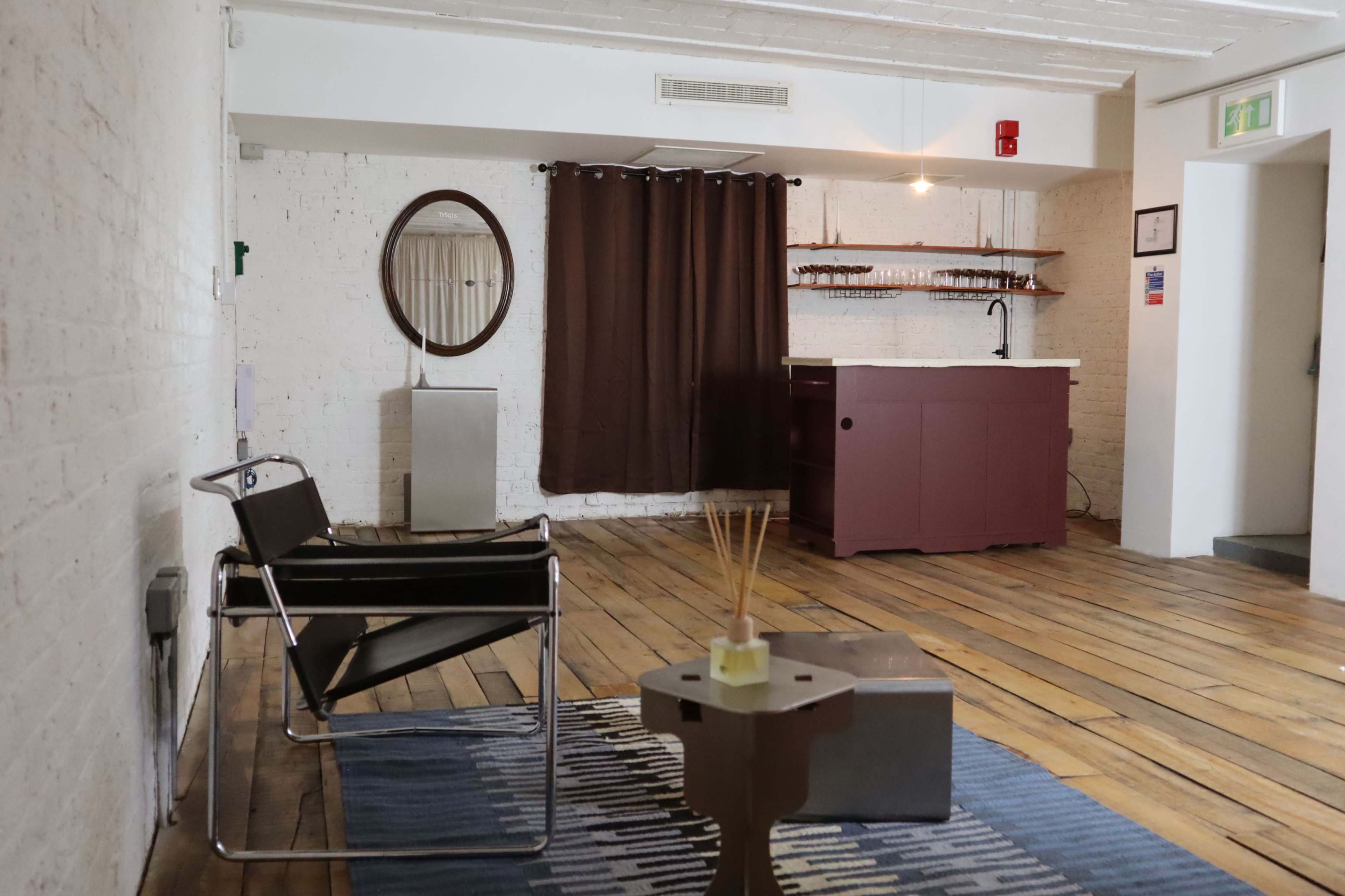 The image shows a minimalistic room with wooden flooring, a black chair, a small side table, a curtain, and a kitchenette area with a sink and countertop.