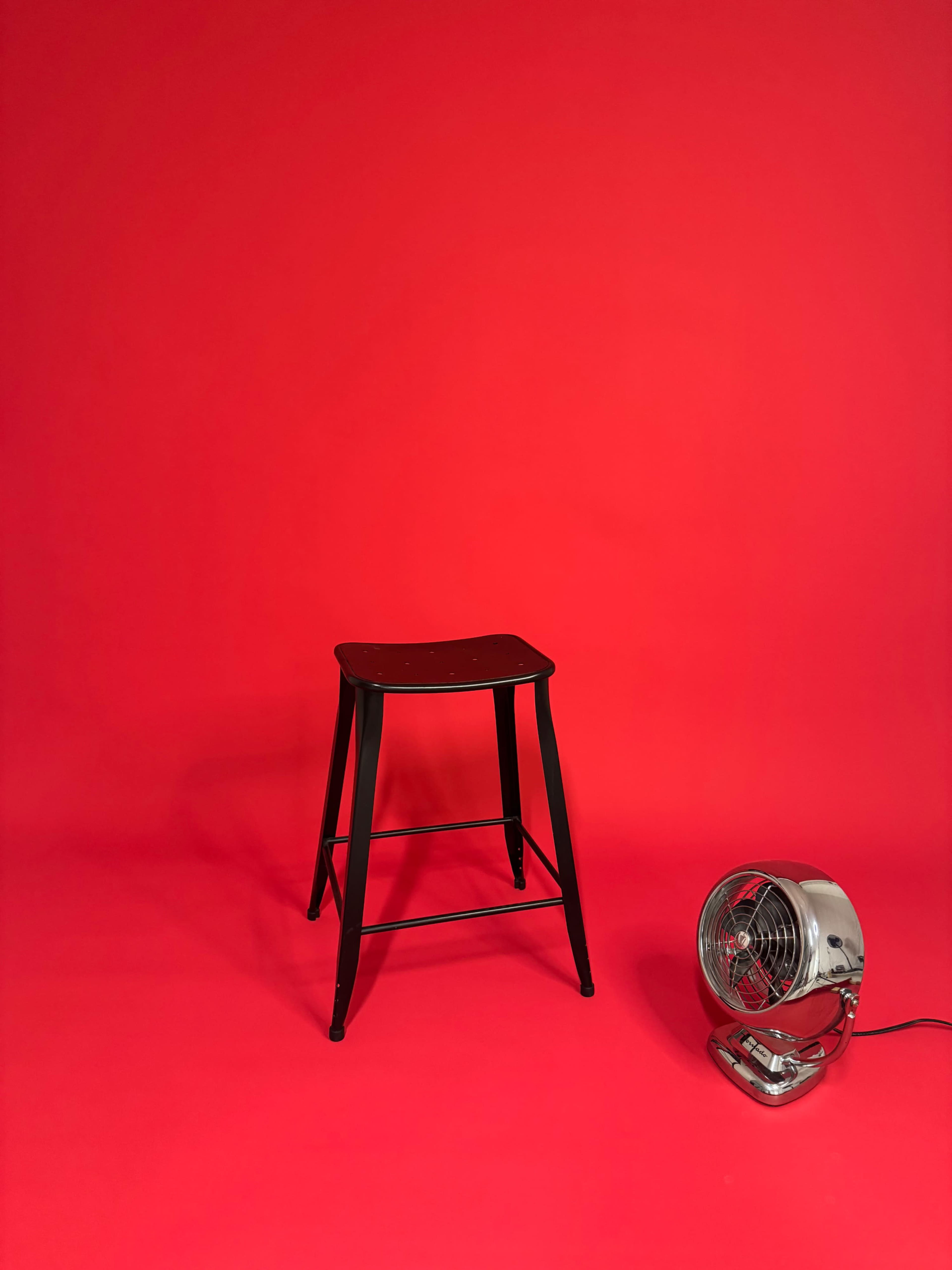 A black metal stool is positioned beside a silver fan against a bright red background.