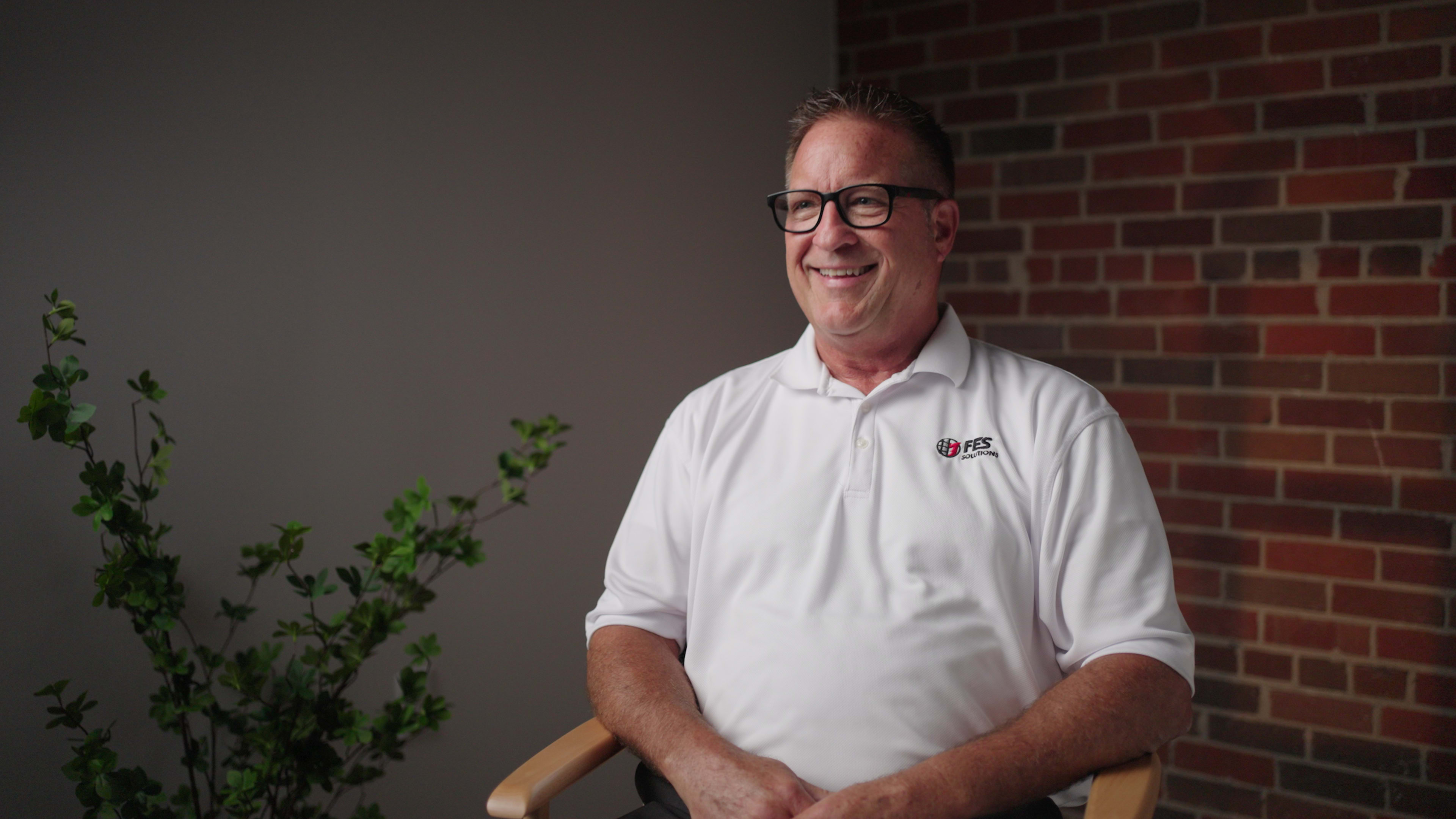 A man in a white polo shirt sits in a chair with a brick wall behind him and a plant nearby.