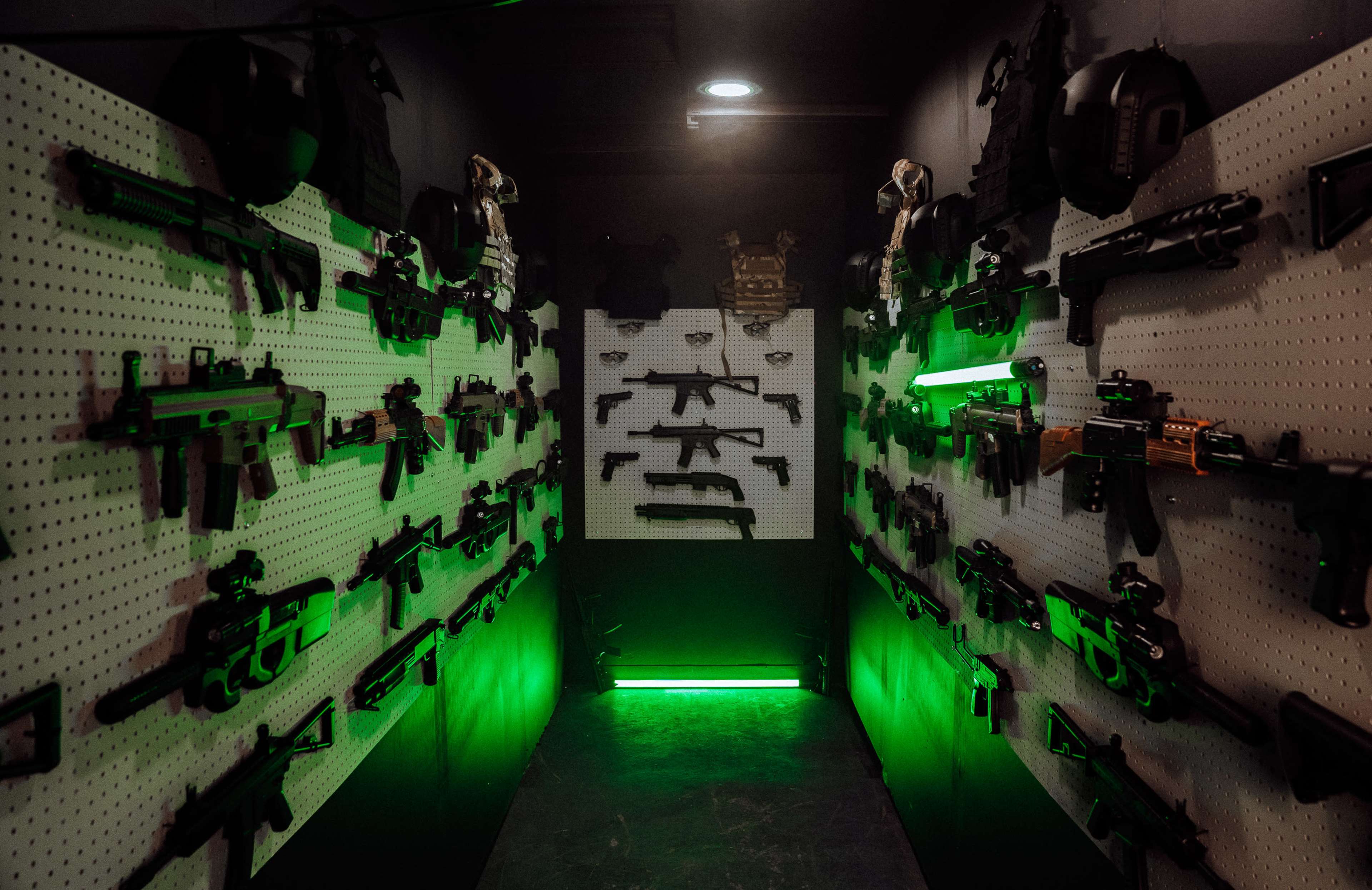 The image shows a narrow room with walls displaying various firearms arranged on pegboards, illuminated by green light.