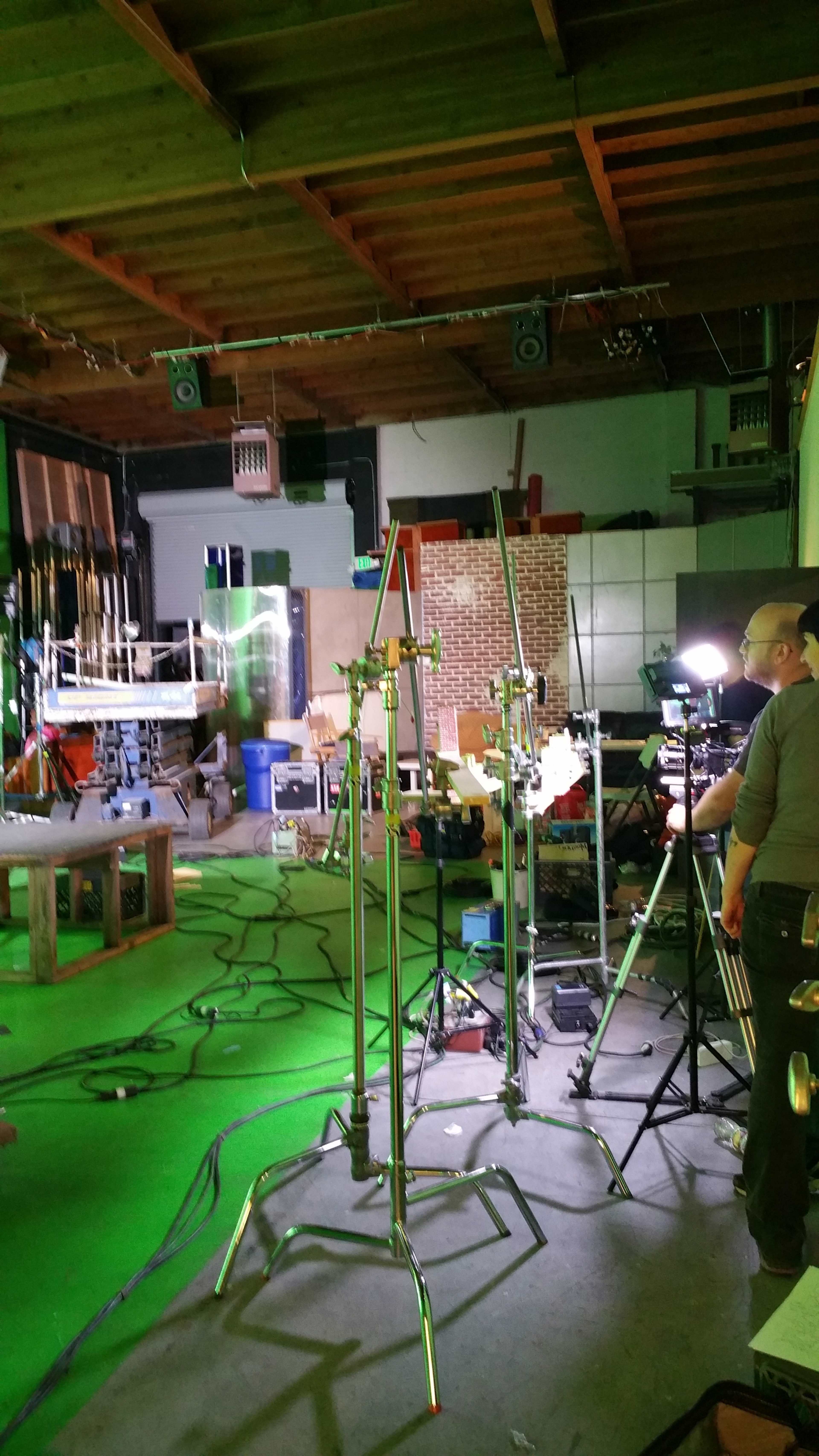 The scene shows a cluttered studio with various equipment, including light stands and cables, against a backdrop of props and sound equipment.