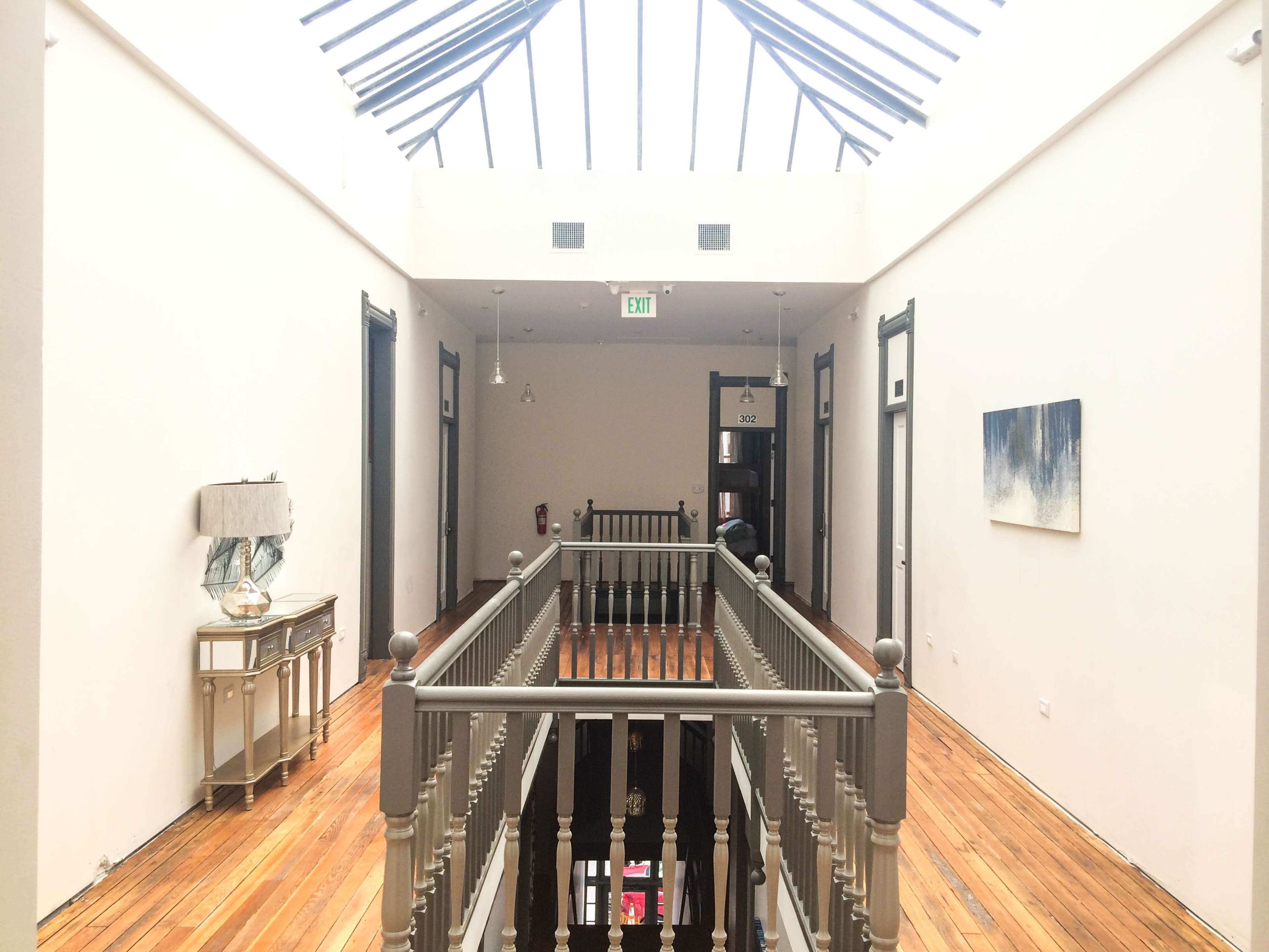 A hallway with wooden floors and a glass skylight features a railing along the upper level, leading to several doors and a piece of artwork on the wall.