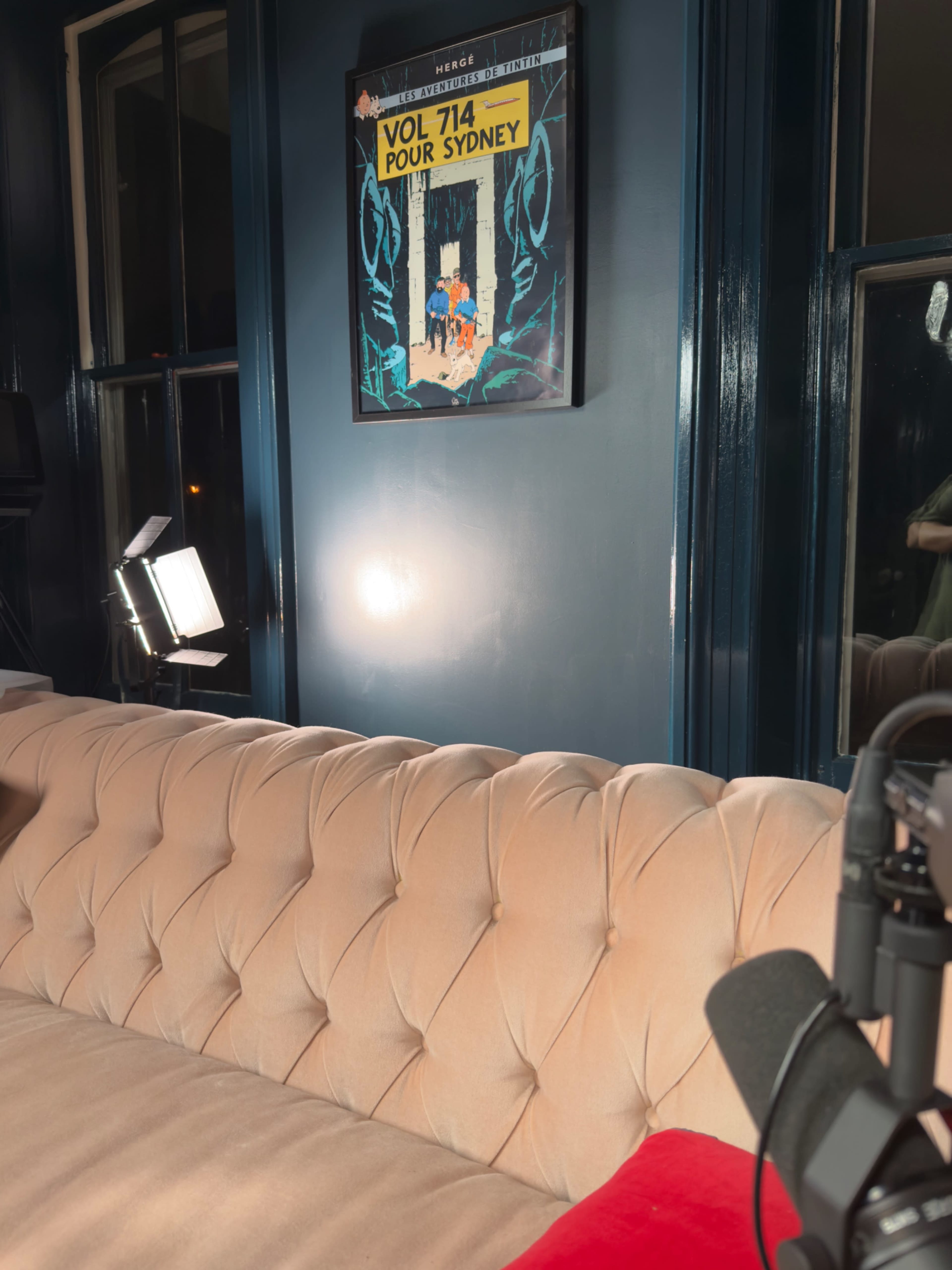 A tufted beige sofa is positioned in a dimly lit room with a poster titled "VOL 714 POUR SYDNEY" hanging on the wall above it.