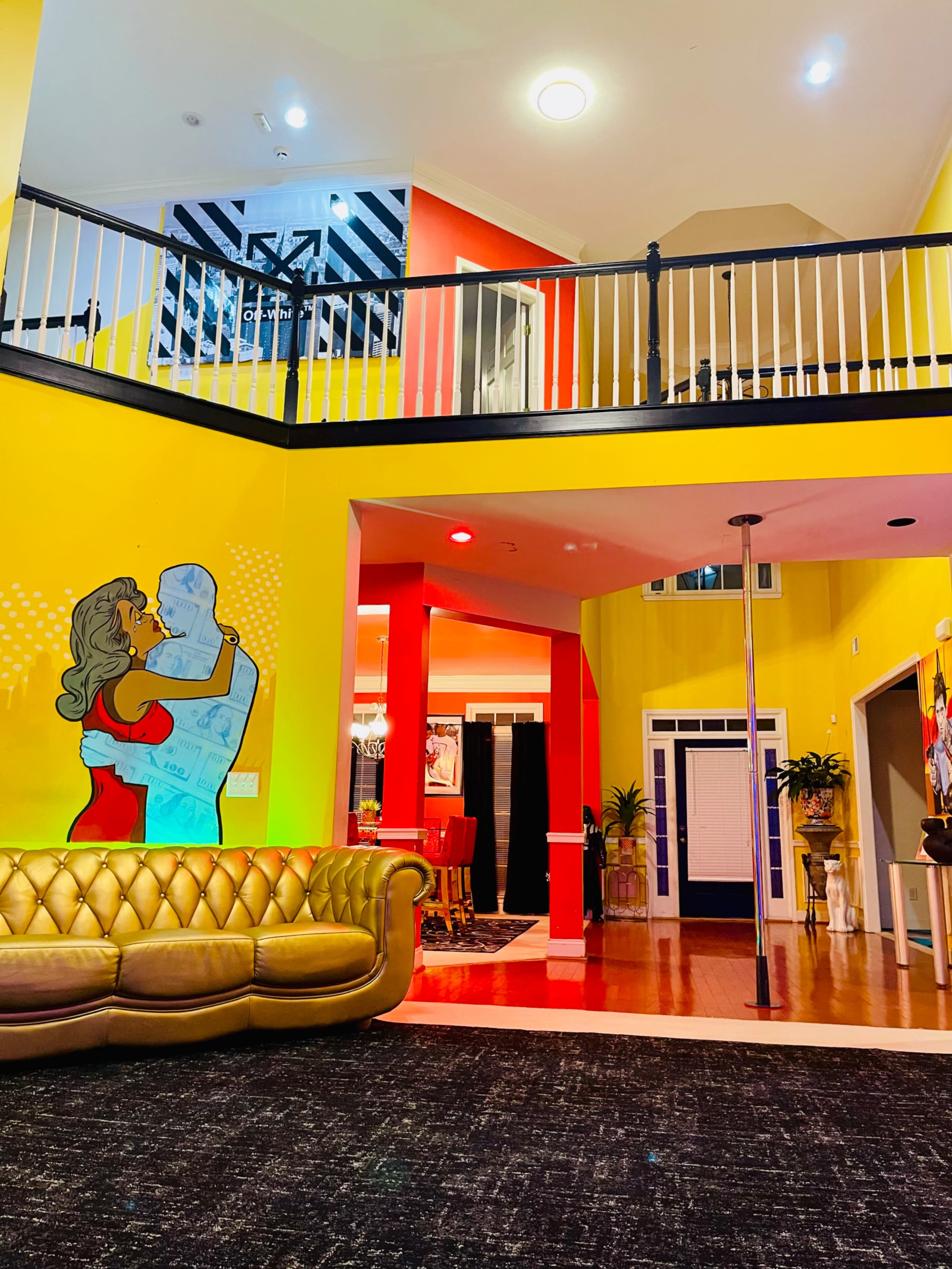 The image shows a brightly colored interior space featuring a large staircase, a bold mural of a couple dancing, and a view of various rooms and furniture, including a yellow couch and a pole.