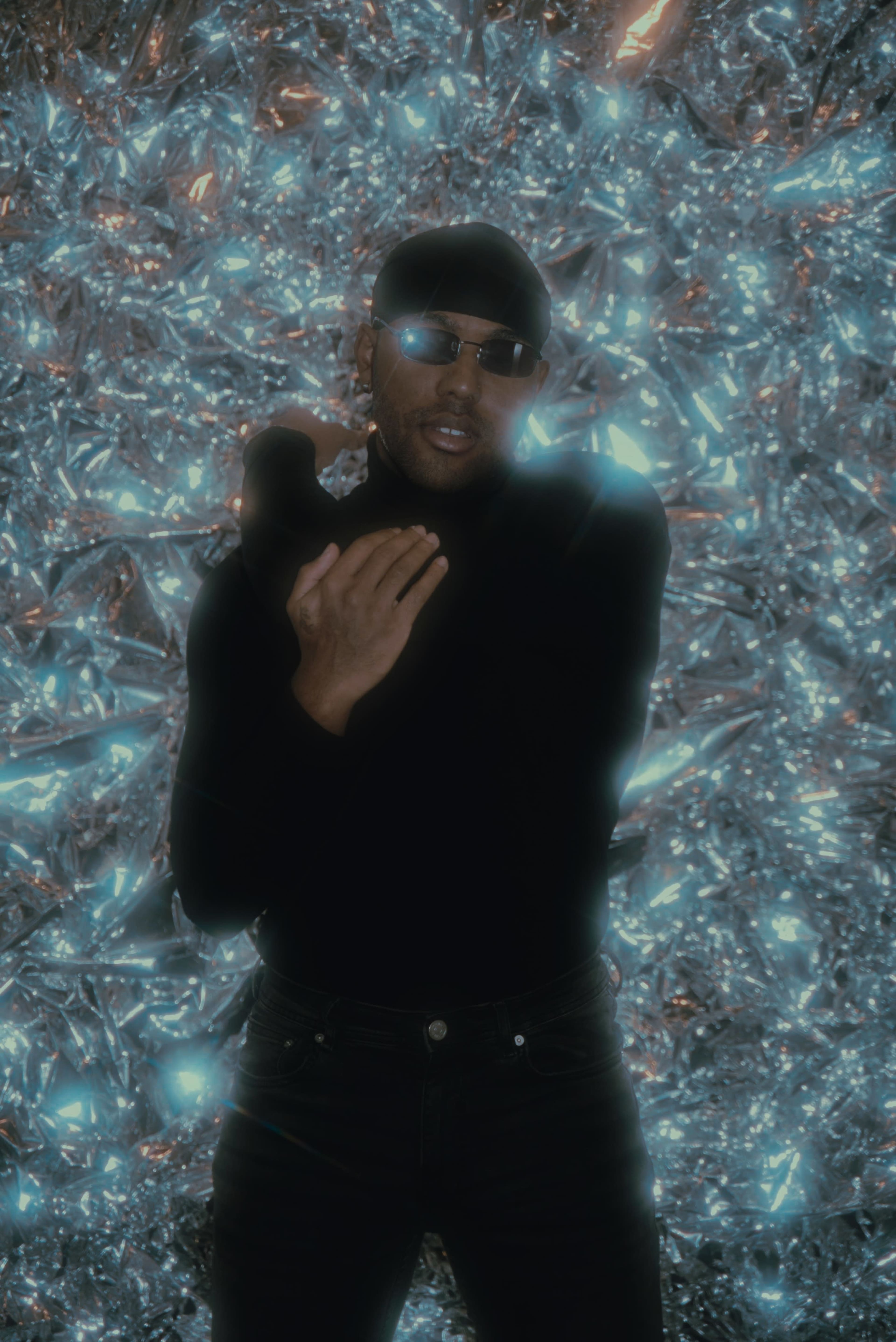 A person wearing a black turtleneck and sunglasses stands against a shimmering, metallic backdrop.
