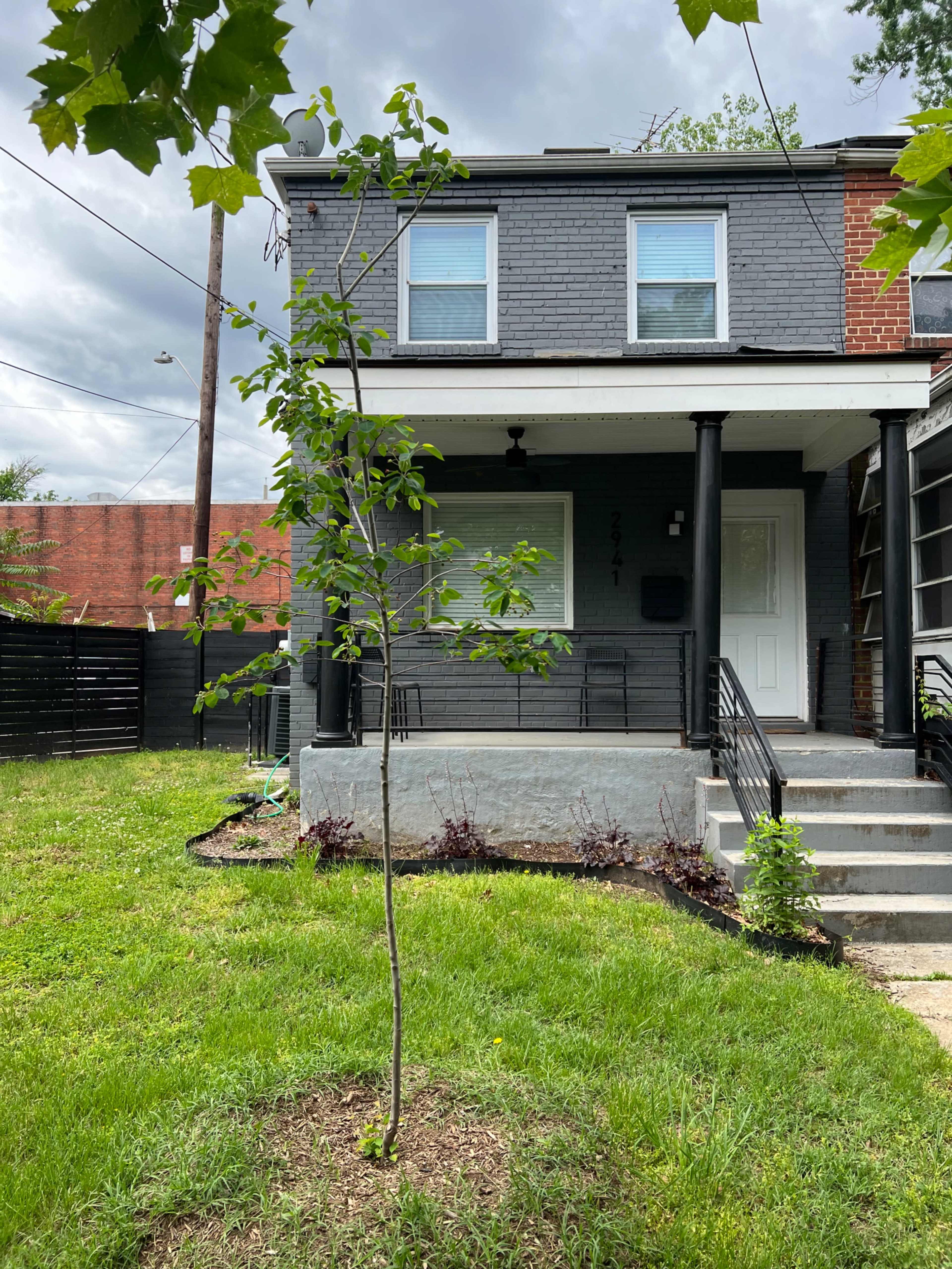 The image shows a two-story gray house with a small tree and grass in the front yard, featuring a porch with stairs and a white door.