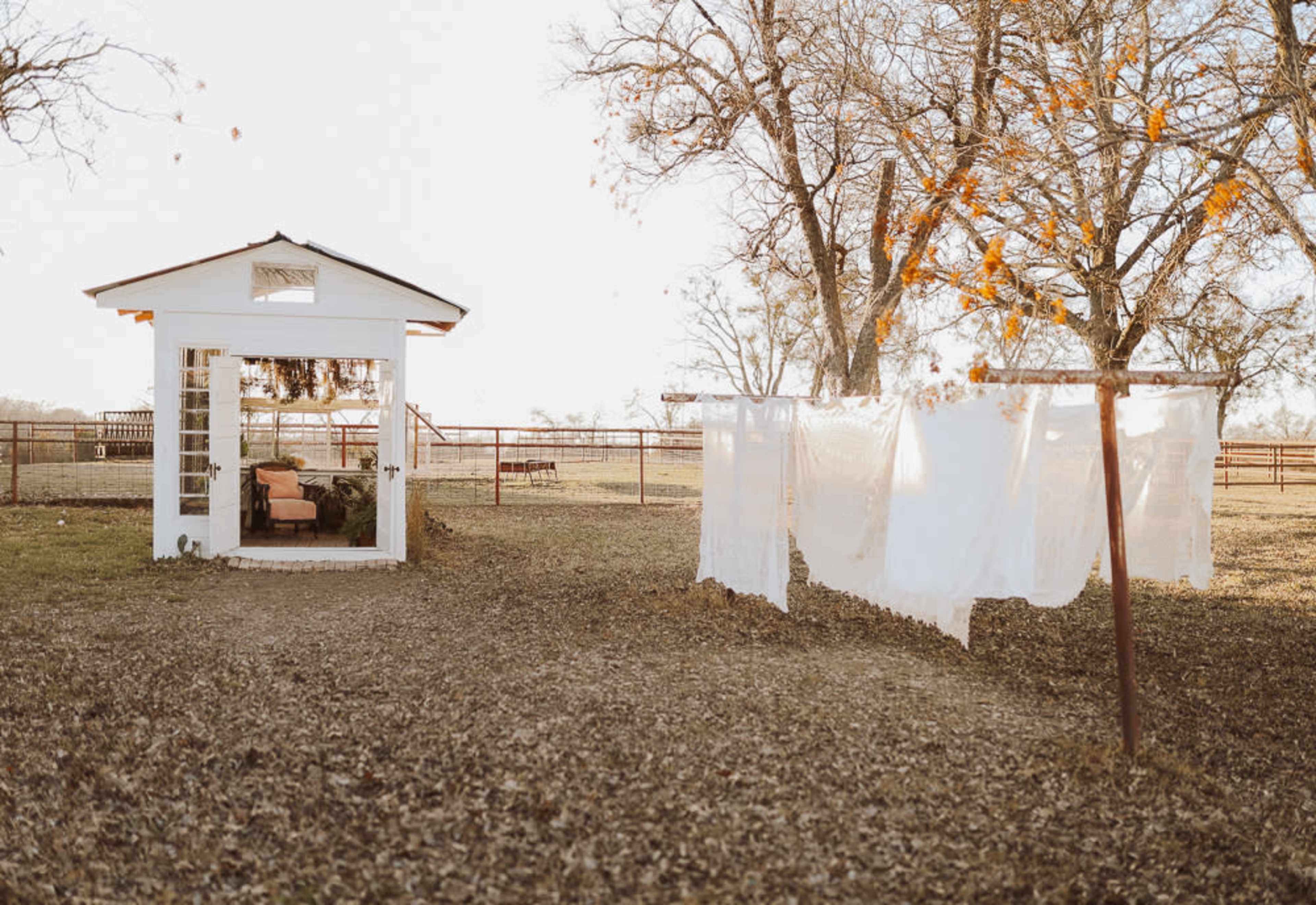 A small white structure with large windows stands beside a clothesline holding freshly laundered white sheets in a grassy area surrounded by trees.