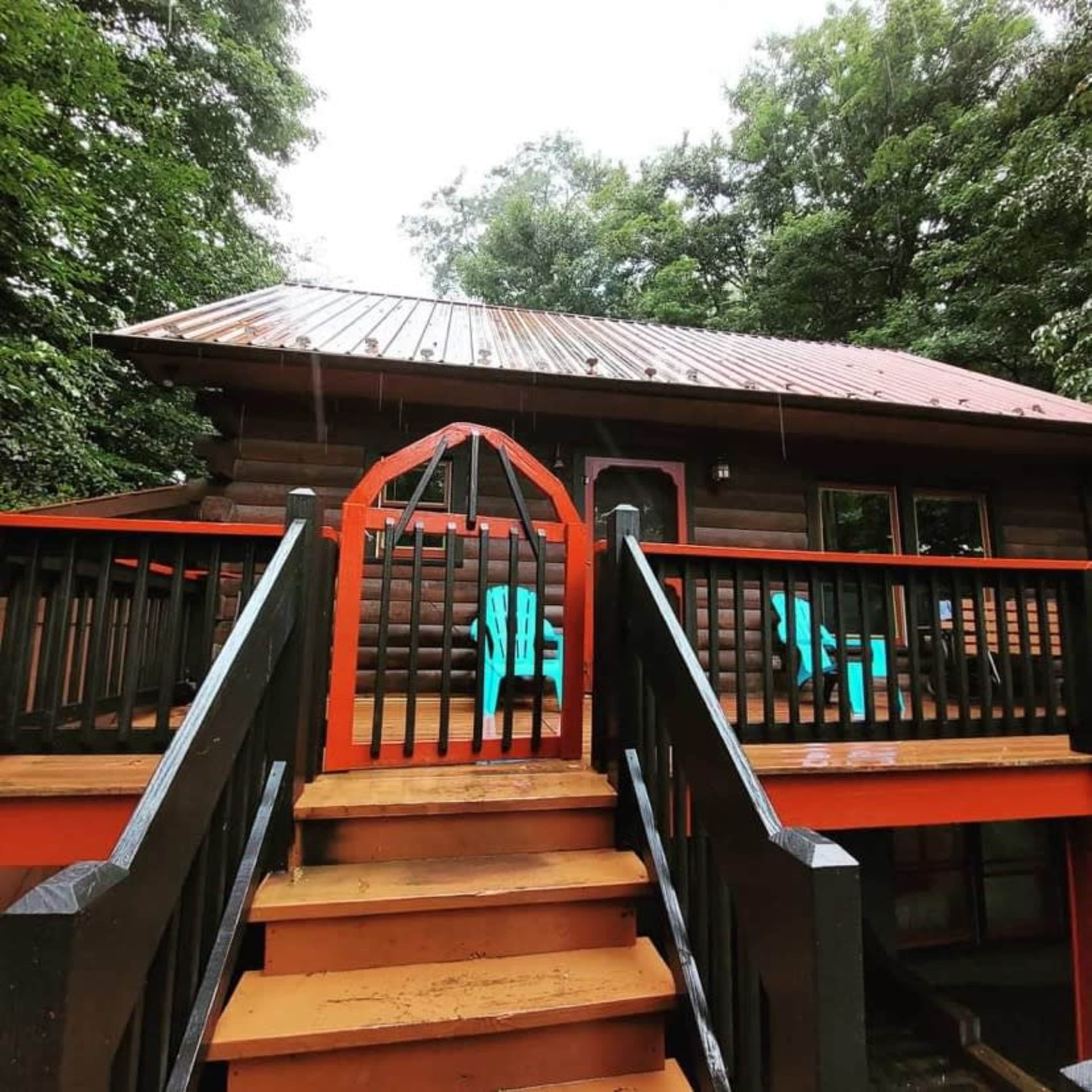 A wooden cabin with a metal roof is situated among trees, featuring a staircase leading up to a deck with brightly colored chairs.