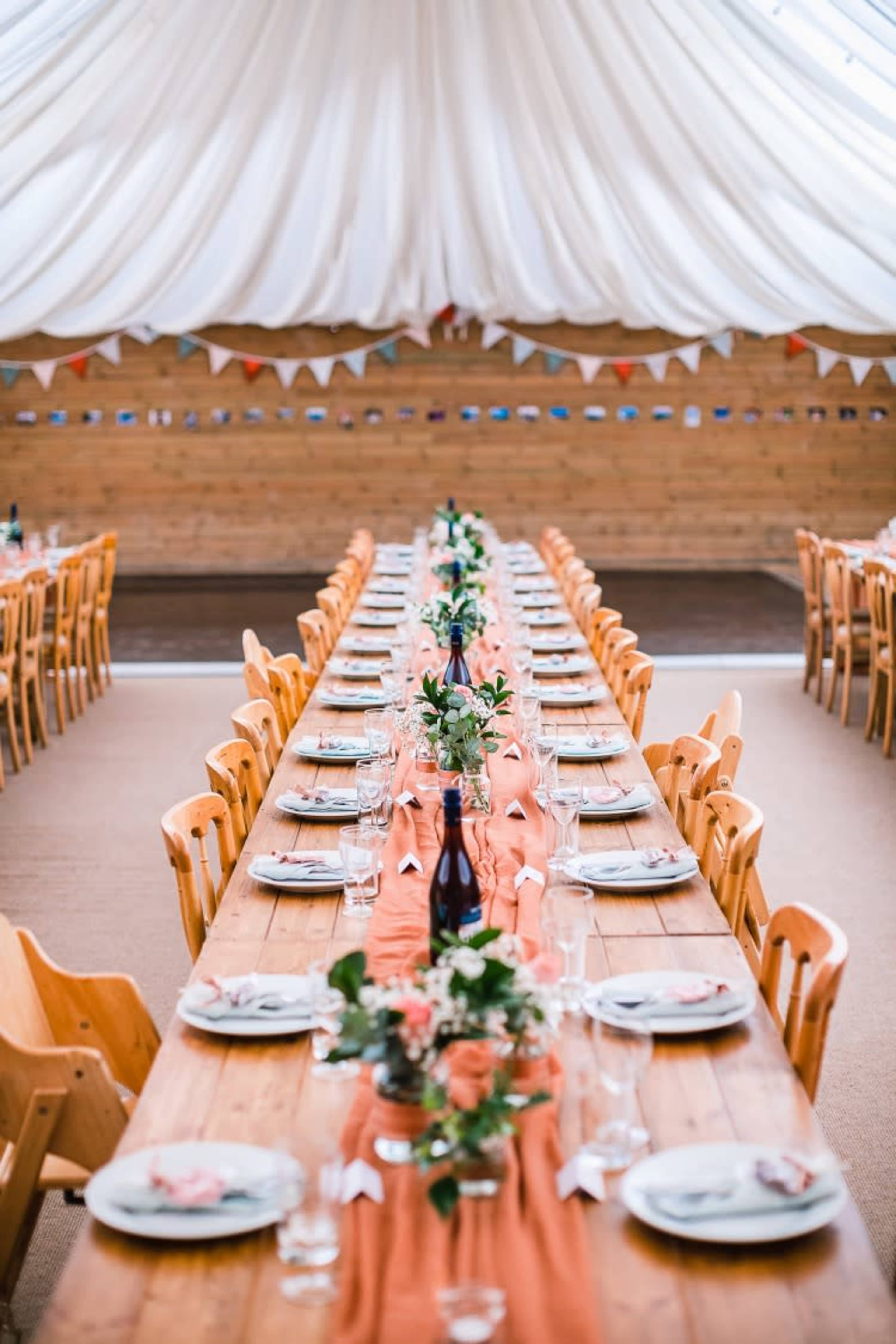 A long banquet table is set with plates and glasses, adorned with flowers, under a white tent with decorative bunting.