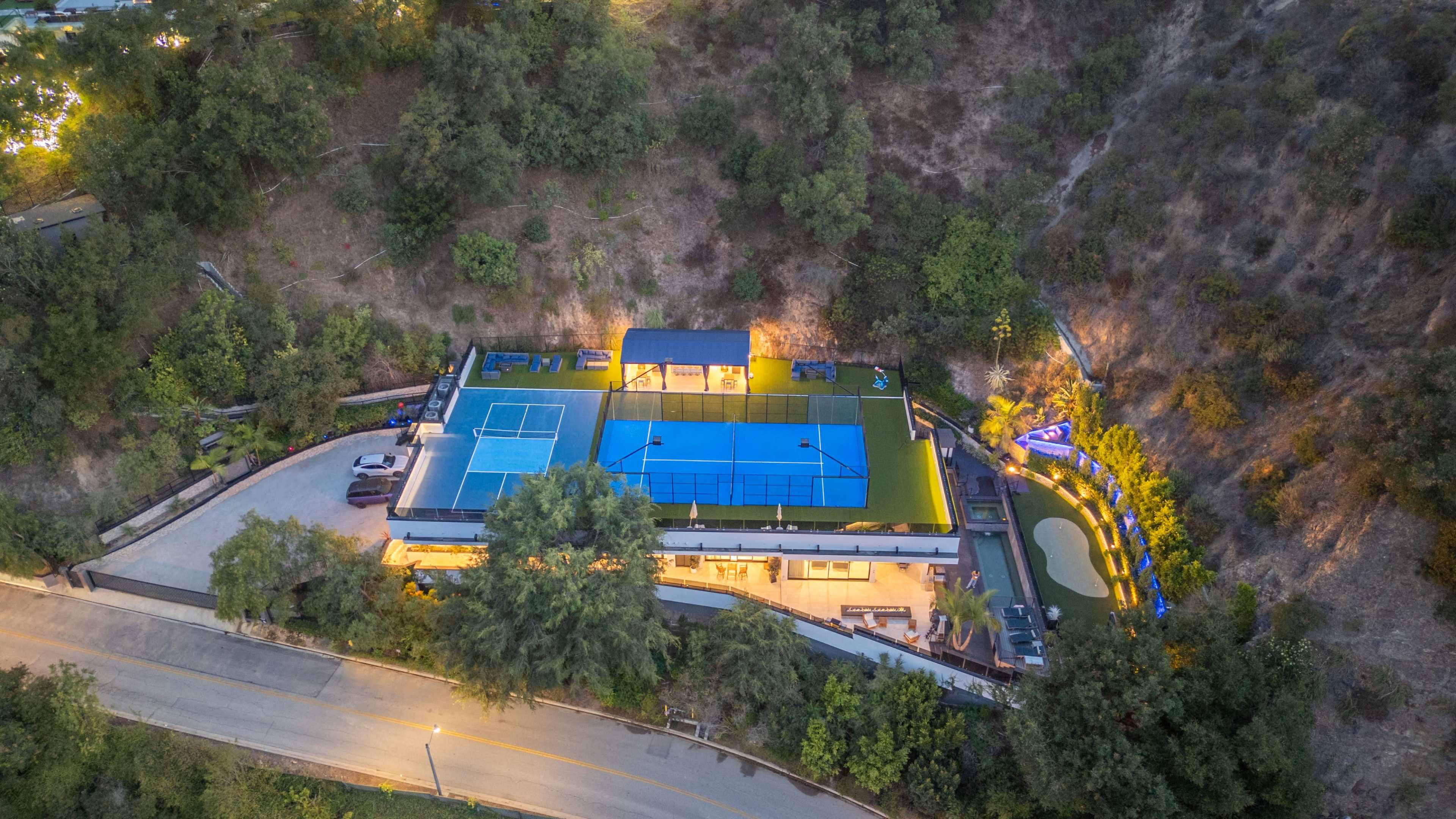 The image shows a modern property featuring two tennis courts, surrounded by lush greenery and a road below.