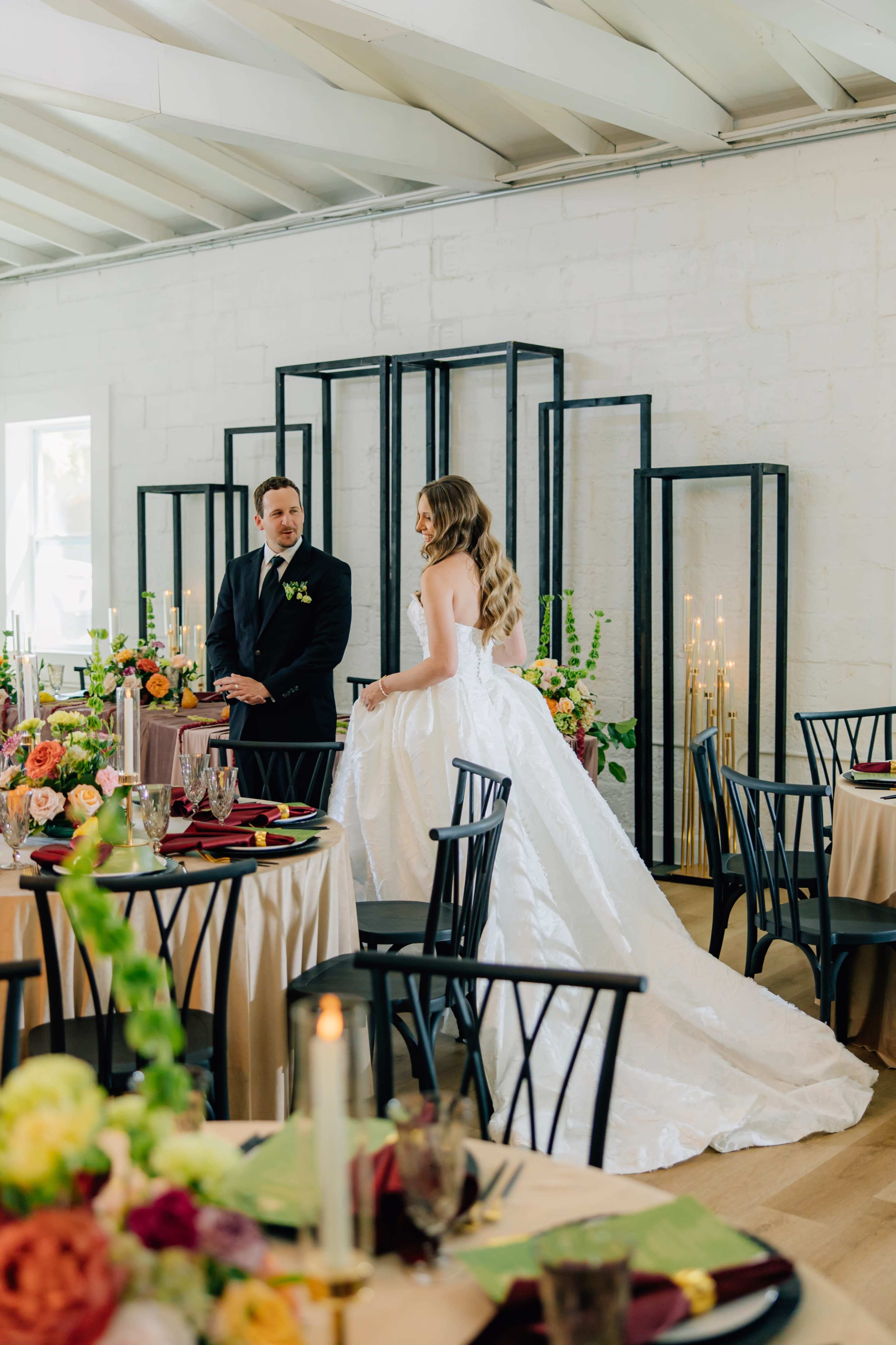 A bride in a white gown stands beside a groom in a black suit, with elegantly set tables and floral arrangements in a well-lit reception venue.