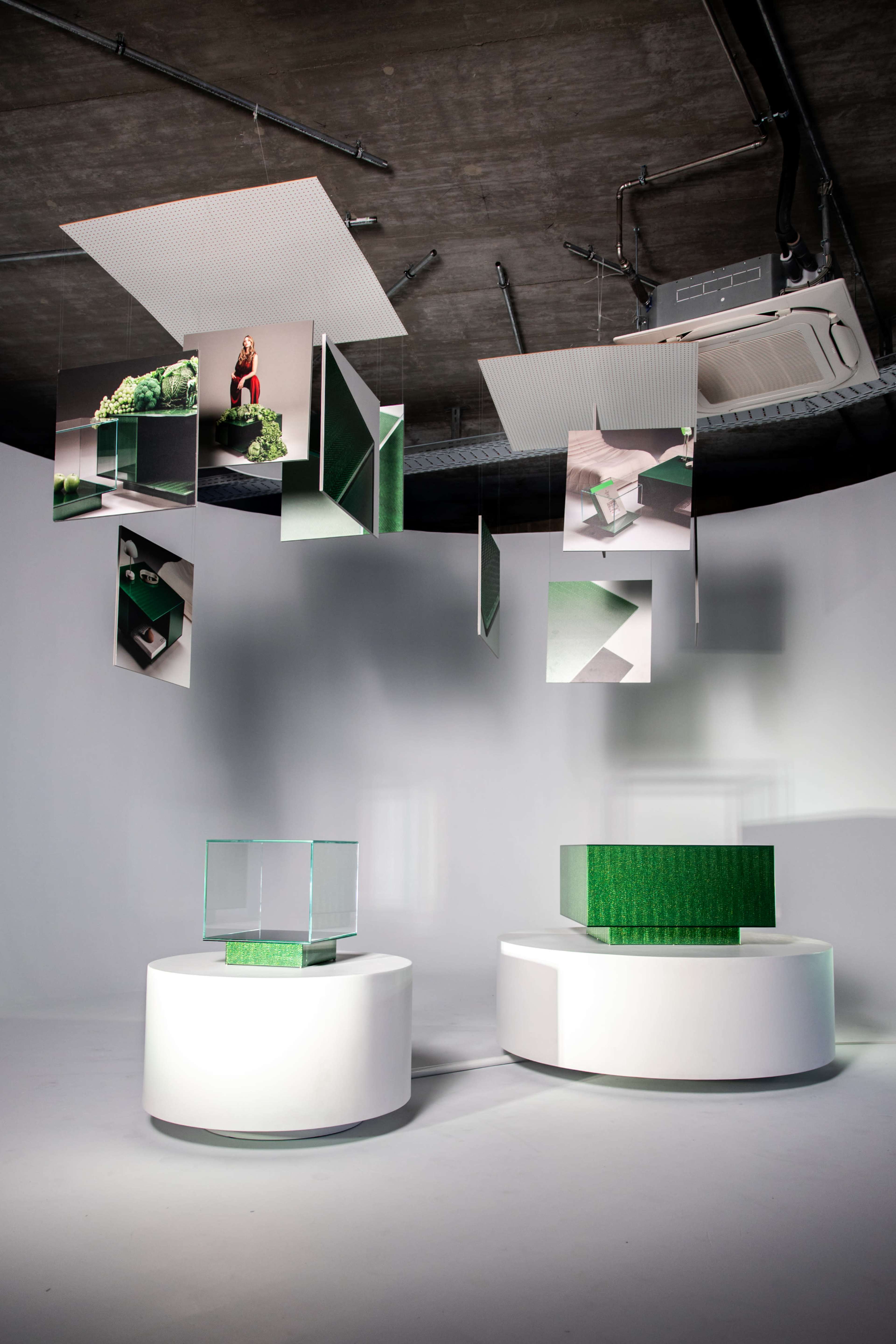 The image features two circular display pedestals in a minimalist setting, with hanging rectangular panels displaying images and green graphic elements above them.