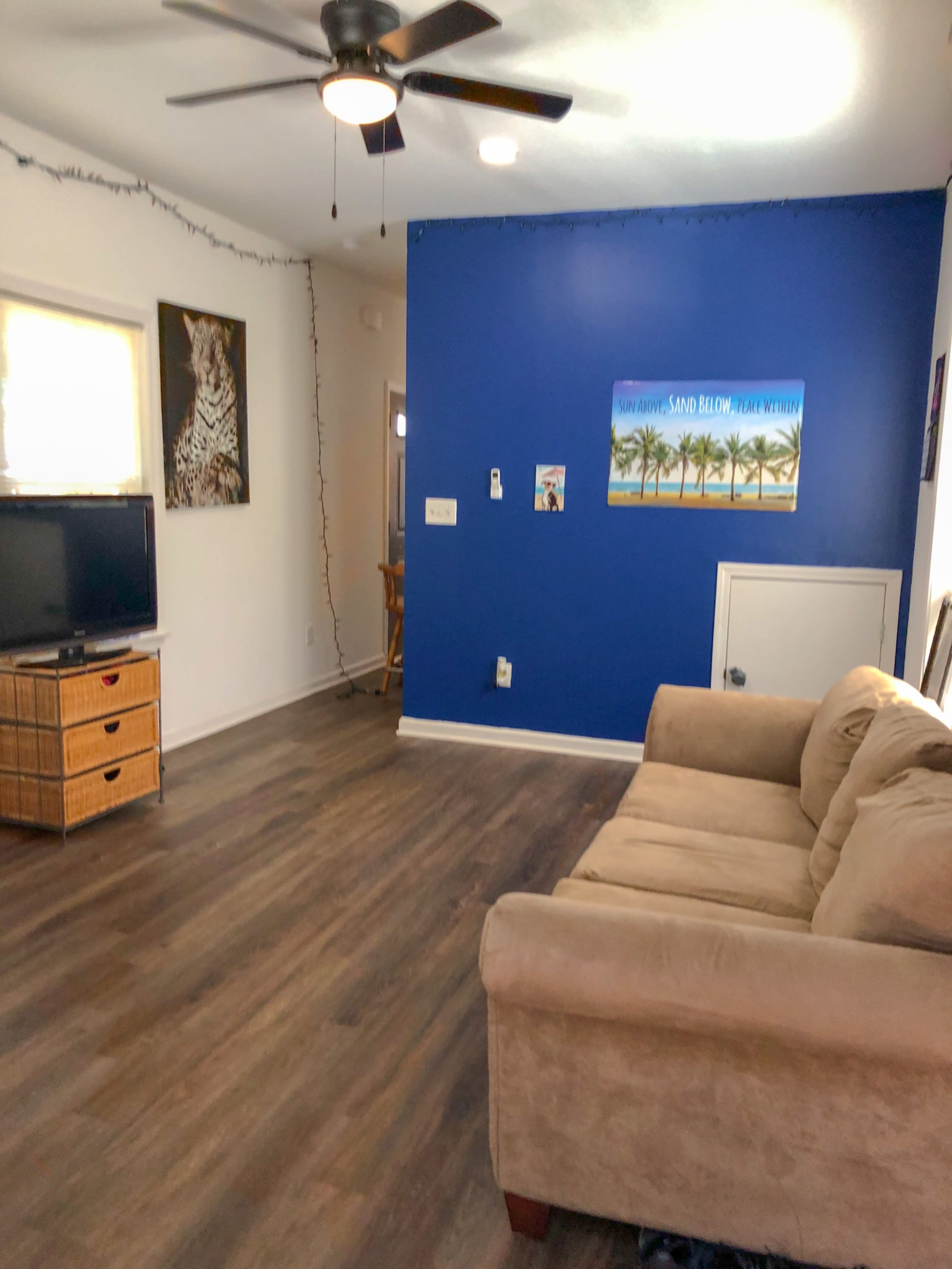 The image shows a living room with a brown couch, a television on a wooden stand, a blue accent wall, and a small dining area visible in the background.